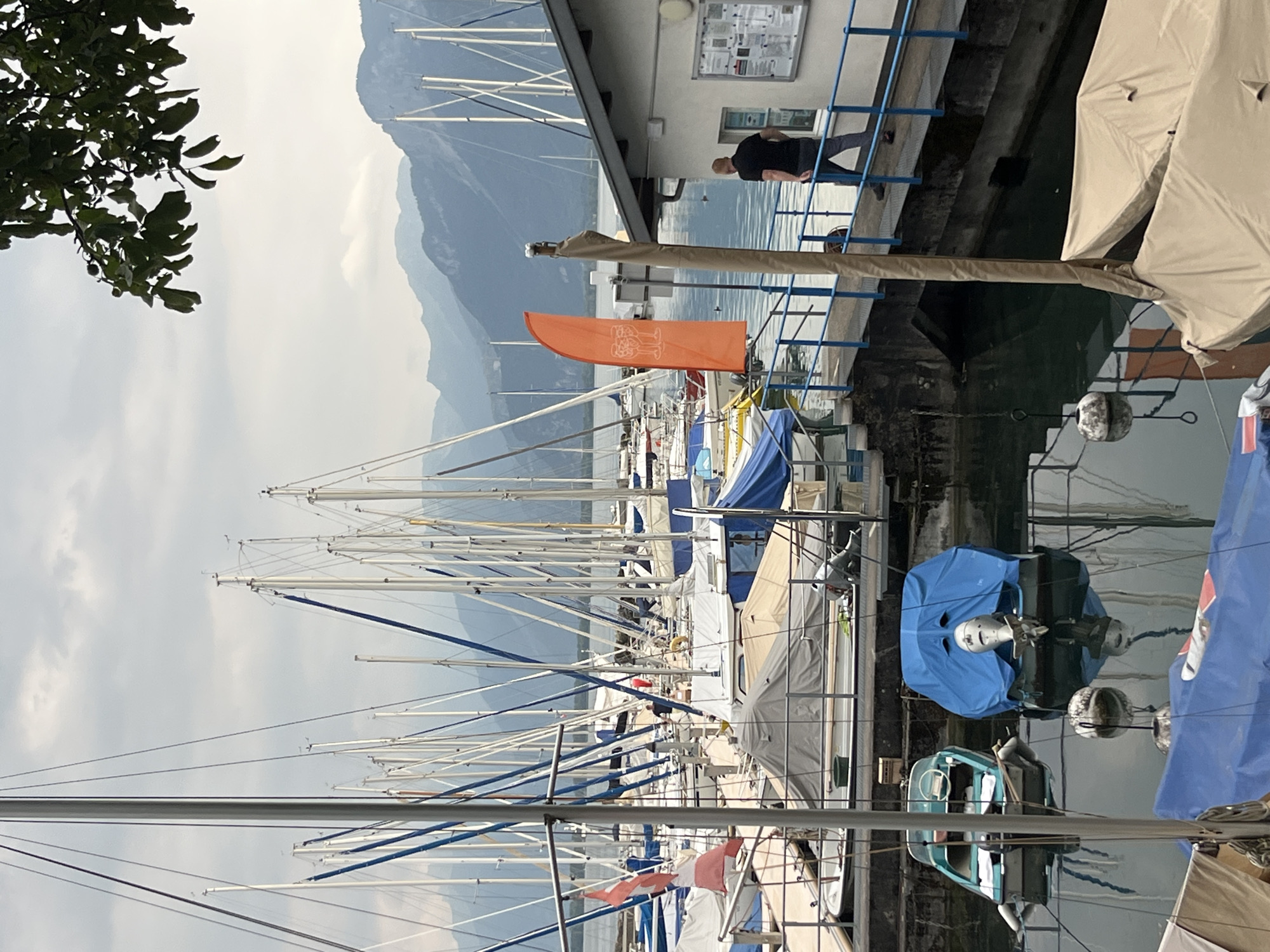 The image shows a marina in Montreux, Switzerland, with numerous sailboats docked. The boats are of various sizes, with many covered with tarpaulins or boat covers. In the foreground, there are several smaller boats, one of which is teal and has a white steering wheel, while another is covered in a blue tarp. A man in a black shirt and grey pants is walking on a pier towards a building. The building appears to be a small office or boathouse, with windows and a bulletin board. An orange flag with an image of two wine glasses is visible on the pier. In the background, a body of water is visible, with mountains in the distance. The sky is overcast with clouds. The overall scene depicts a peaceful day at a Swiss marina.