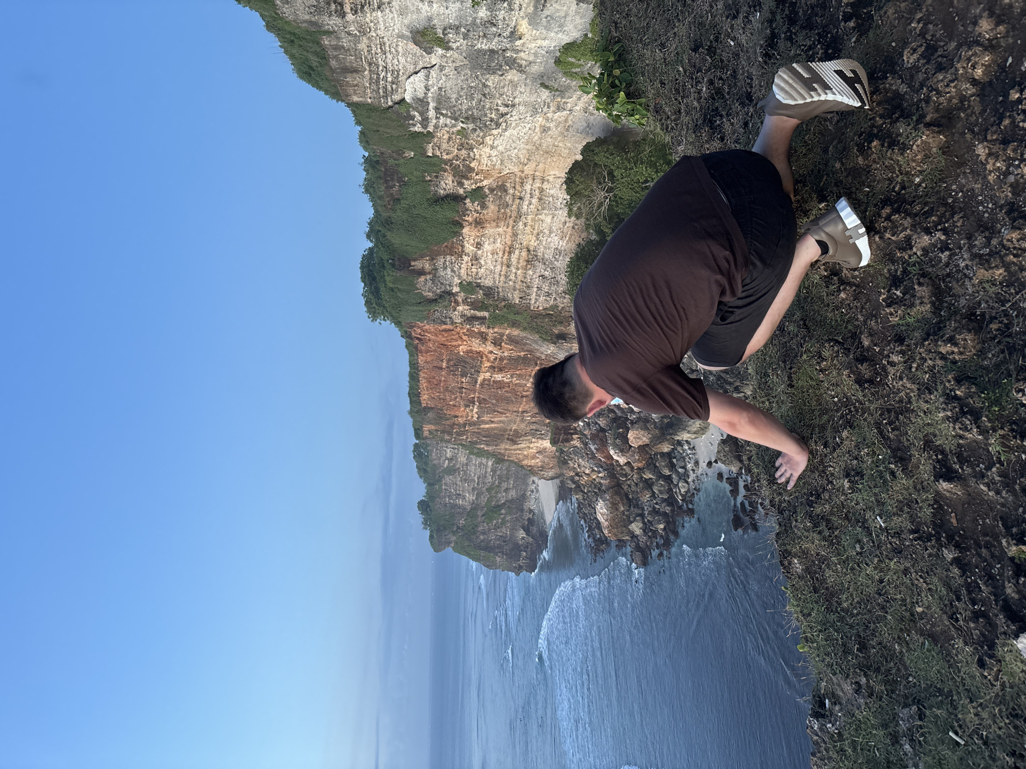 A man leans over the edge of a cliff in Kuta Selatan, Indonesia. The cliff is tall, with visible layers of rock and vegetation. The man is wearing a brown t-shirt, black shorts, and light-colored sneakers. Below the cliff, waves crash against rocks and a small beach. The sky is clear and blue.