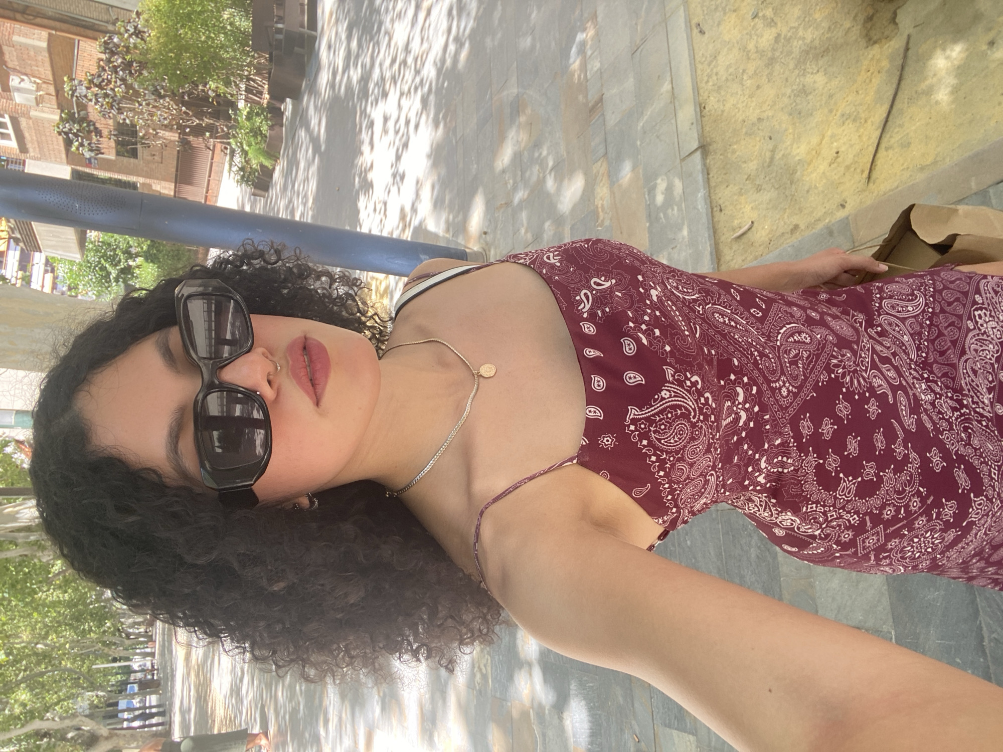 A young woman with dark, curly hair is taking a selfie outdoors in a city. She is wearing oversized black sunglasses, a thin silver chain necklace with a gold pendant, and a burgundy dress with a white paisley pattern. She has a nose ring. In her right hand, she is holding a brown paper bag.
The background suggests an urban environment with trees lining a walkway. The walkway is paved with stone tiles, and there is a blue pole to the right of the woman. Buildings and more greenery are visible in the distance. The lighting indicates it is daytime, with dappled sunlight on the ground.
The scene is consistent with a street or park area within a city. While the specific location is not definitively identifiable from the image alone, the presence of Mediterranean vegetation, architecture, and the general ambiance could be characteristic of a city like Murcia, Spain, particularly its tree-lined promenades or plazas.