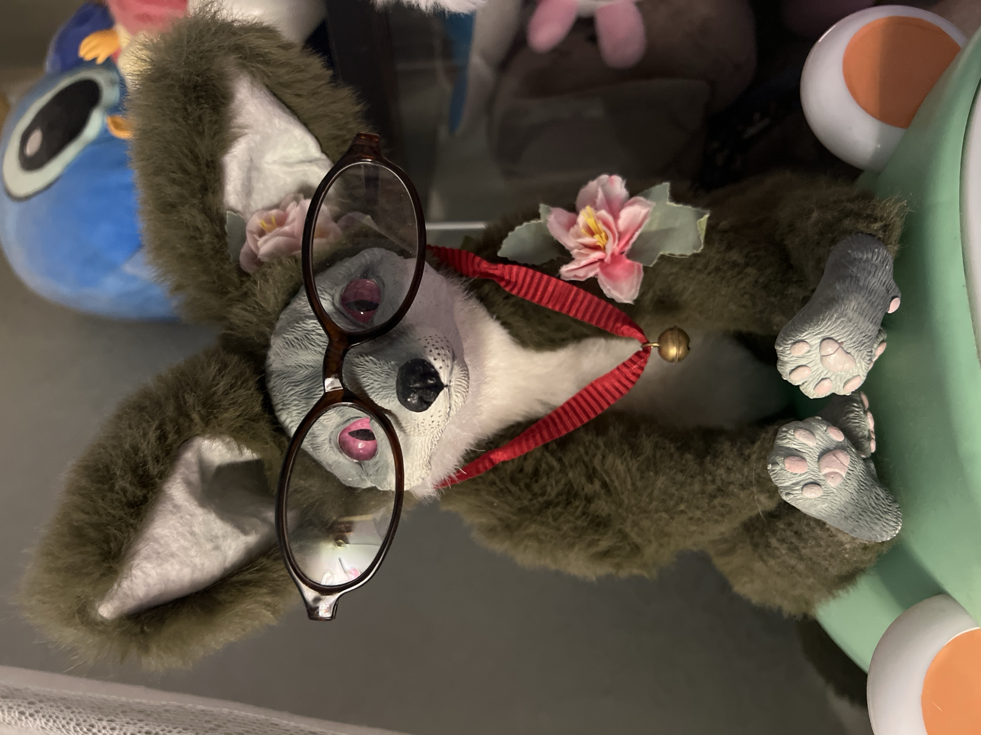 A stuffed animal, resembling a fox with large, fluffy ears and pink eyes, sits wearing glasses and a red ribbon with a bell and flower. The stuffed animal is positioned in the foreground. In the background, other stuffed animals and toys are visible, including a large blue stuffed character with black eyes. The stuffed animal is resting on a light green surface. The overall scene appears to be an indoor setting, possibly a shelf or display area. There is no discernible information in the image that would indicate its location in Moscow, Russia.