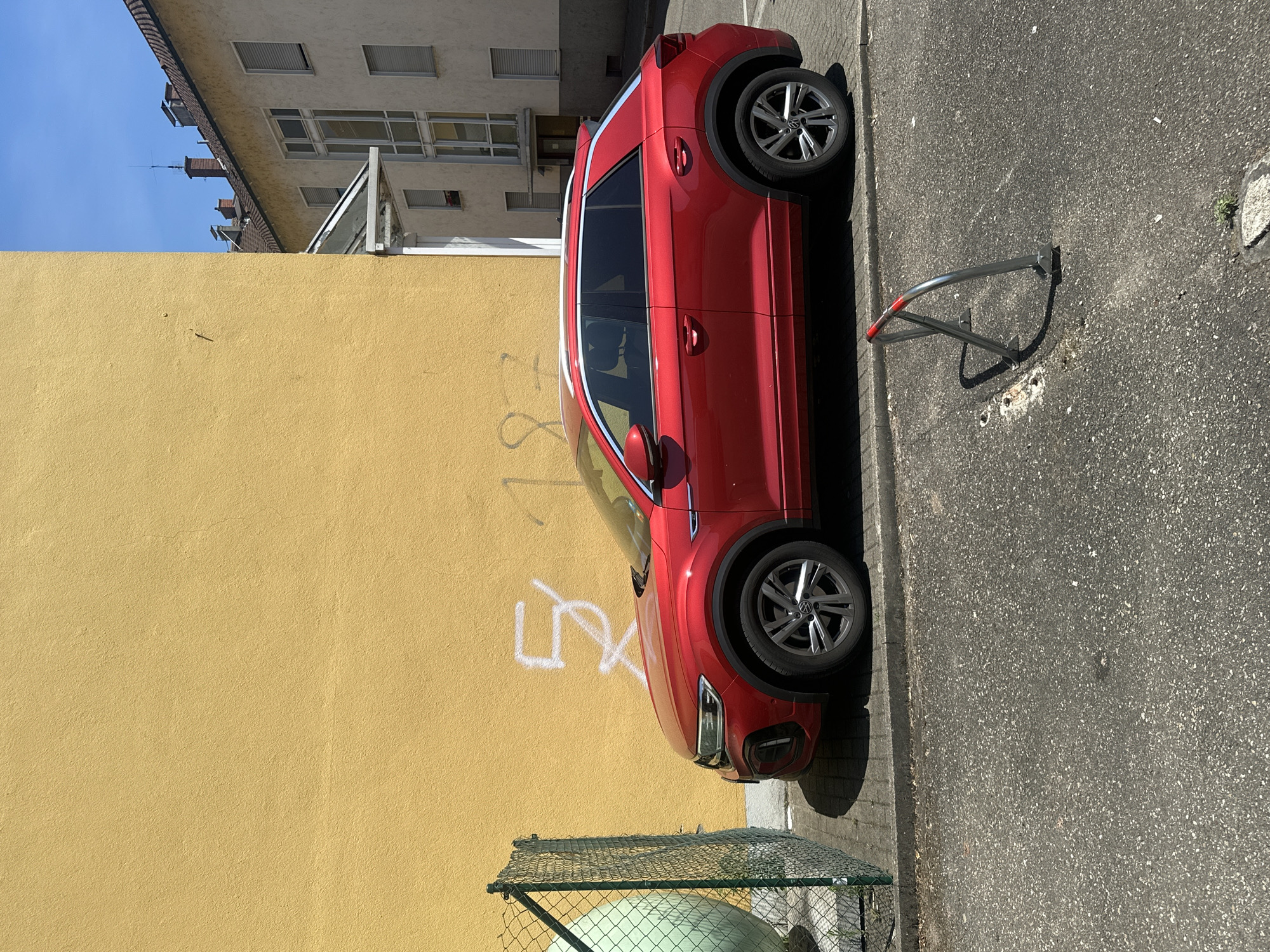 A red Volkswagen T-Roc SUV is parked on asphalt next to a yellow building and a chain-link fence. The building's facade has white graffiti that appears to spell out "5X" and "187". A metal bike rack is visible in the foreground. The sky is blue with a few clouds. The scene appears to be in an urban environment.