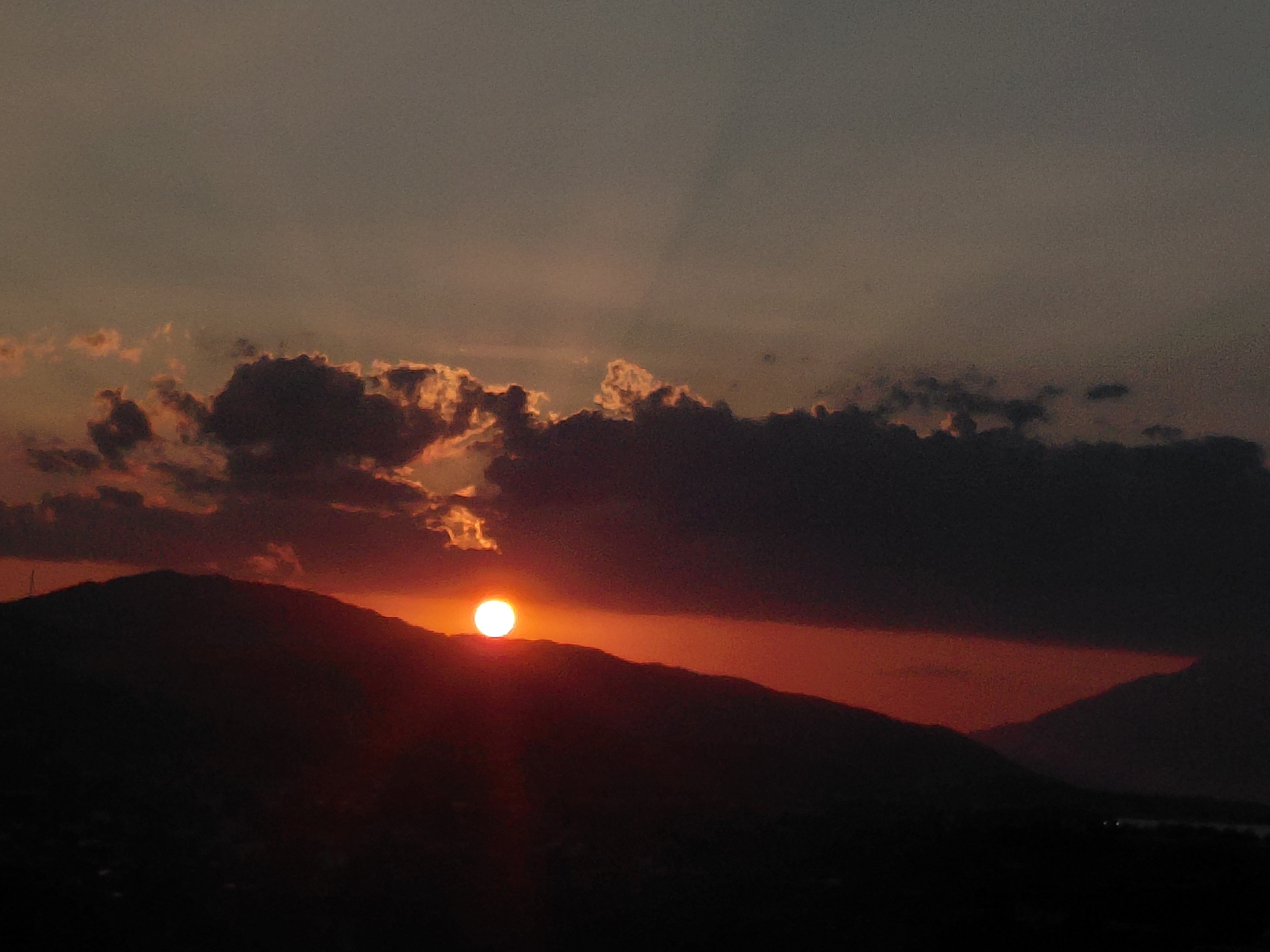 A wide-angle shot captures a sunset over the Municipal Unit of Aegio, Greece, casting a dark silhouette of mountains against a sky ablaze with warm hues. The sun, a brilliant orange orb, is partially obscured by the rugged peaks, its light creating a dramatic halo. Fluffy clouds, tinged with gold and crimson, drift across the horizon, adding texture and depth to the celestial canvas. The foreground is dominated by the dark, undulating forms of mountains, their slopes descending into shadow. In the distance, a hint of water is visible, reflecting the fading light of the day. The overall impression is one of serene natural beauty, a tranquil close to the day.