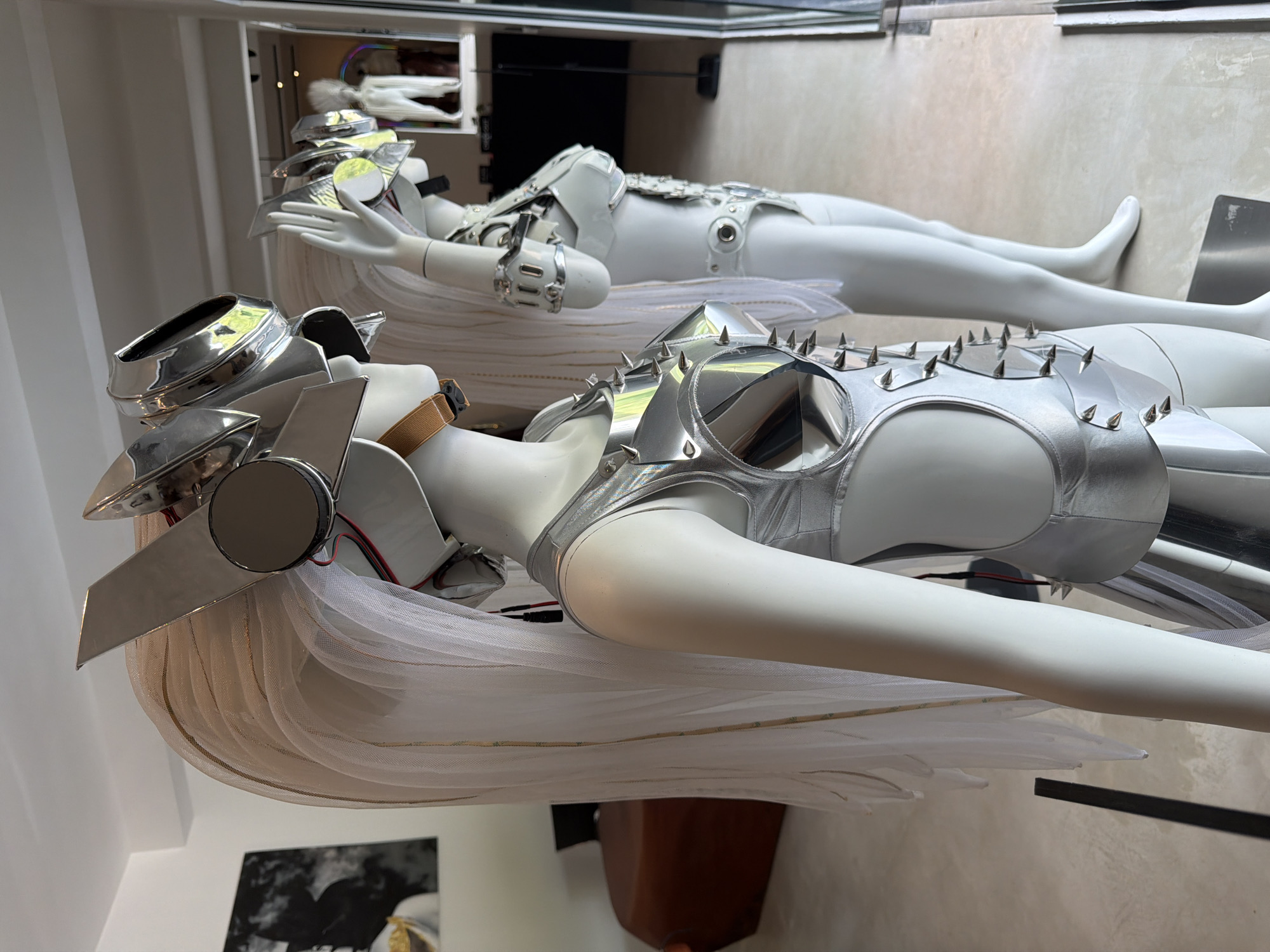 A close-up, medium shot captures two mannequins dressed in elaborate, futuristic silver costumes, displayed indoors. The mannequin in the foreground, viewed from the side and slightly from the front, wears a metallic silver top with angular, spiked embellishments across the chest and a matching skirt. A translucent white veil, adorned with thin gold threads, cascades from its head, framing a metallic, angular helmet with a circular reflective visor.
The second mannequin, positioned behind and to the right of the first, is also clad in a silver costume that appears to be a metallic bra-like top and bikini-style bottoms, also featuring metallic accents. Its helmet is similarly elaborate and reflective. Both mannequins have white, smooth skin and wear white wigs.
The background reveals a brightly lit interior with a polished concrete floor. To the left, a dark, abstract painting is mounted on a white wall. Further in the background, beyond the mannequins, a retail-like environment is visible, with additional displays and figures that are out of focus. The overall impression is that of a fashion exhibit or a retail display in a modern establishment. The specific location of Ko Samui District, Thailand, is not evident from the visual cues in the image itself.