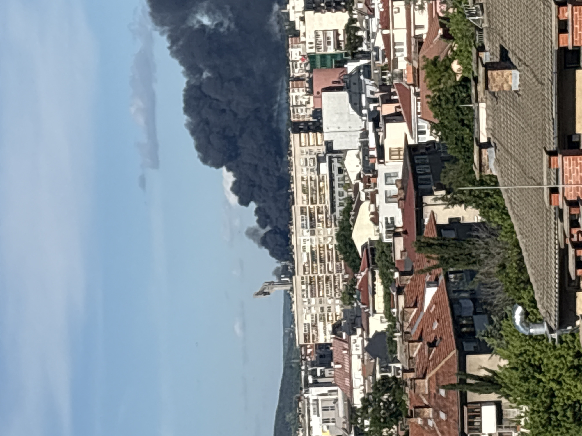 The image shows a cityscape, likely taken from an elevated position. Rows of buildings with varying heights and roof colors (predominantly red and white) stretch into the distance. Dominating the left side of the image is a large plume of dark smoke that obscures the sky. A body of water is visible in the distance. Given the prompt location of Stara Zagora, Bulgaria, it is likely that the scene is somewhere in that city. The source of the smoke plume is not identifiable.