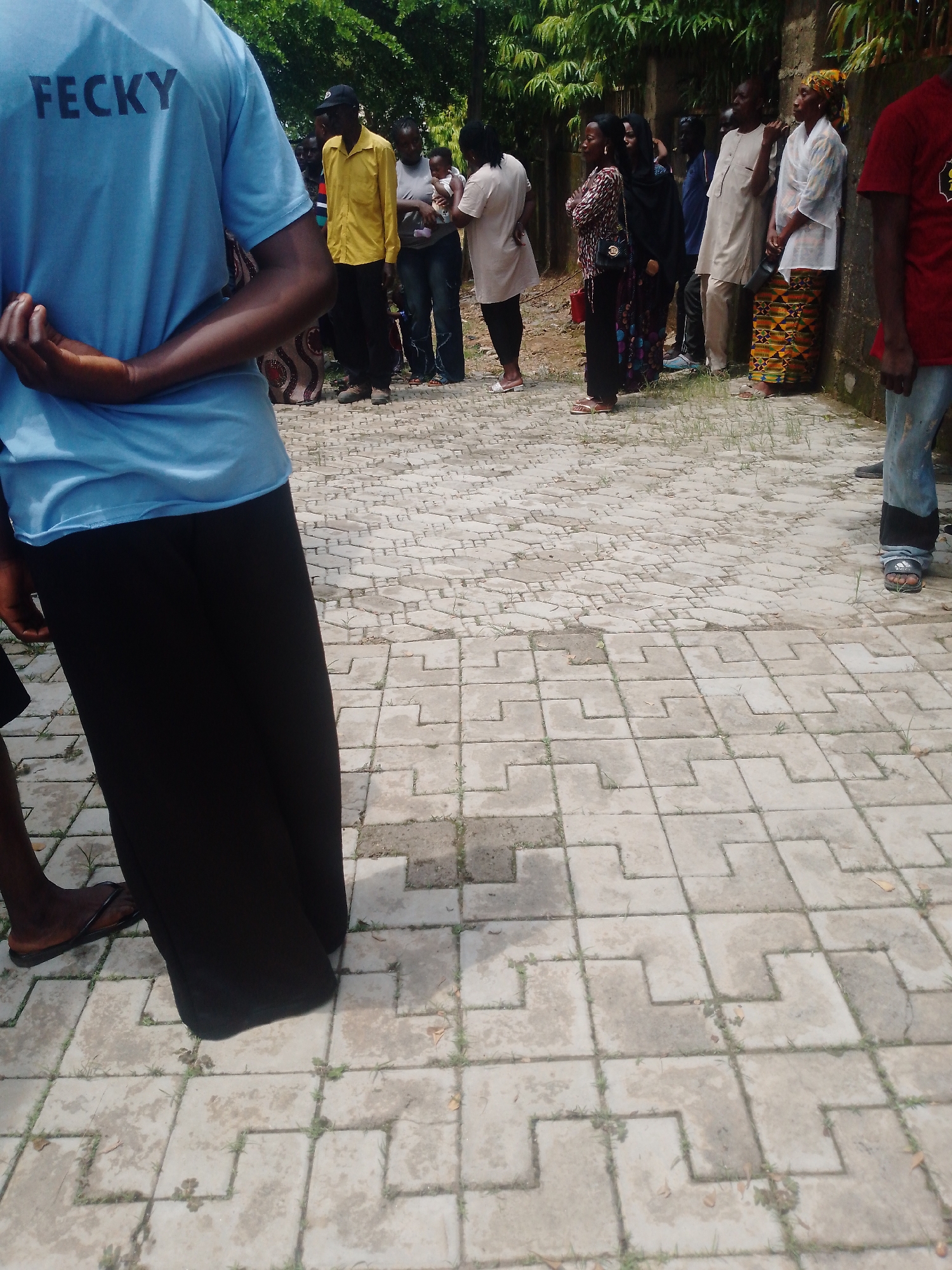 In Akwanga, Nigeria, a group of people stands in a line on a paved walkway. The individual in the foreground, from the waist down, wears a light blue t-shirt with the word "FECKY" printed in black on the back and a long, black skirt. Their hands are clasped behind their back. To their right, several other individuals are visible. A person in a bright yellow shirt and black pants stands next to a woman holding a baby. Further down the line, other men and women are dressed in various traditional Nigerian attire, including a man in a beige outfit and a woman in a colorful skirt and white headscarf. The ground is covered with interlocking hexagonal concrete pavers, with sparse green grass growing in the cracks. In the background, a wall and dense green foliage can be seen.