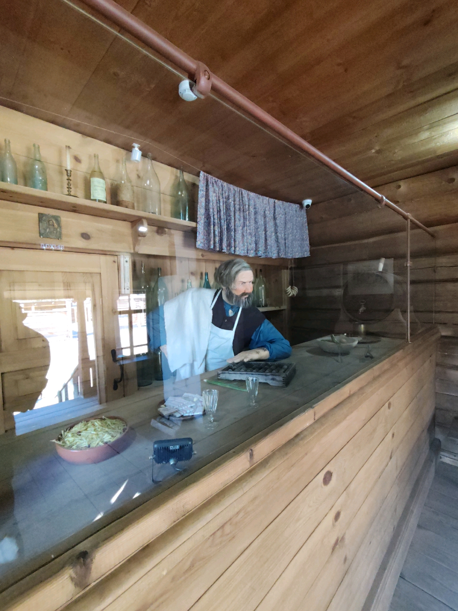 The image shows a diorama inside a wooden building, likely part of the "Taltsy" museum complex in Irkutsk, Russia. A mannequin dressed in period clothing, representing a man with a beard and wearing an apron over a blue shirt, stands behind a wooden counter. He is depicted with his hands resting on an abacus and appears to be engaged in some transaction or calculation.
On the counter in front of the mannequin are several items: a glass of liquid, a stack of papers, and what appears to be a small mortar and pestle. To the left of the mannequin, a clay dish filled with what looks like chopped straw or hay is visible.
Behind the counter, a shelf displays various old bottles, a candelabra, and a small framed image. A patterned fabric curtain hangs from the wall above the shelf. Two security cameras are mounted on the ceiling. The environment is constructed of rough-hewn wooden logs and planks, characteristic of historical buildings in the region. The scene is viewed through a glass barrier, which causes some reflections. The lighting appears to be a mix of ambient light and possibly interior lighting.
