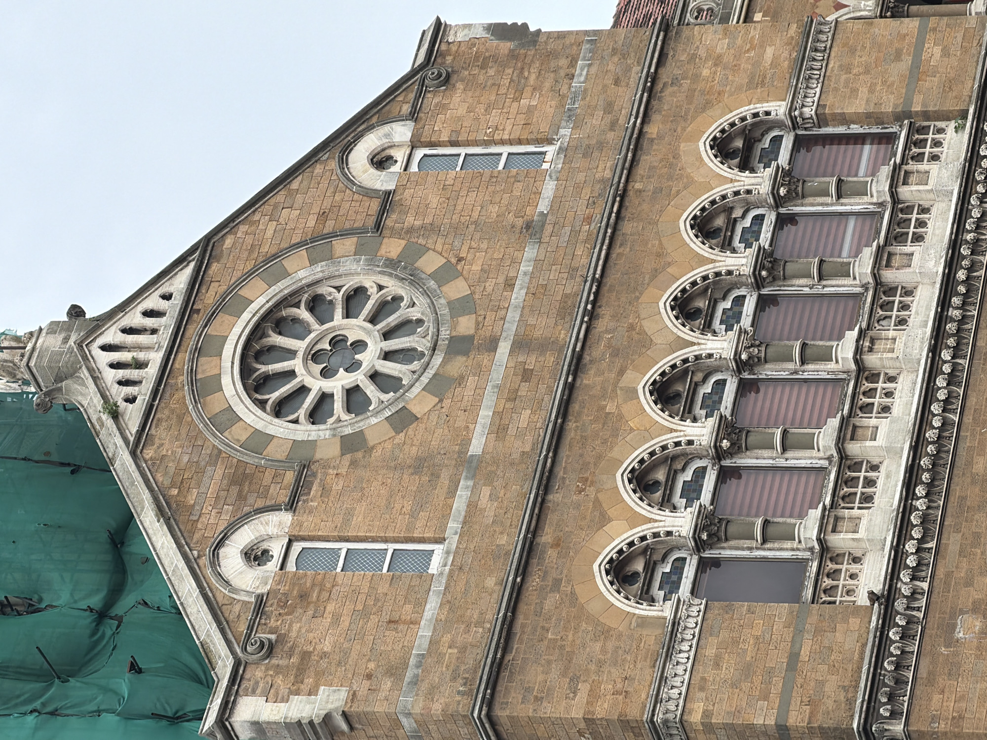 The image shows a detailed view of the exterior of the Chhatrapati Shivaji Maharaj Terminus (CSMT) in Mumbai, India. The architecture is Gothic Revival, characterized by the intricate stonework, arched windows, and the prominent rose window at the top. The building is constructed with warm-toned sandstone. A green tarpaulin covers some upper portions of the building, suggesting ongoing construction or renovation. Below the rose window, a row of arched windows with ornate columns and decorative grilles is visible. A small bird is perched on the decorative ledge below these windows. The sky is overcast and a pale blue-gray. The image is a close-up, focusing on the architectural details of the upper facade of the historic railway station.