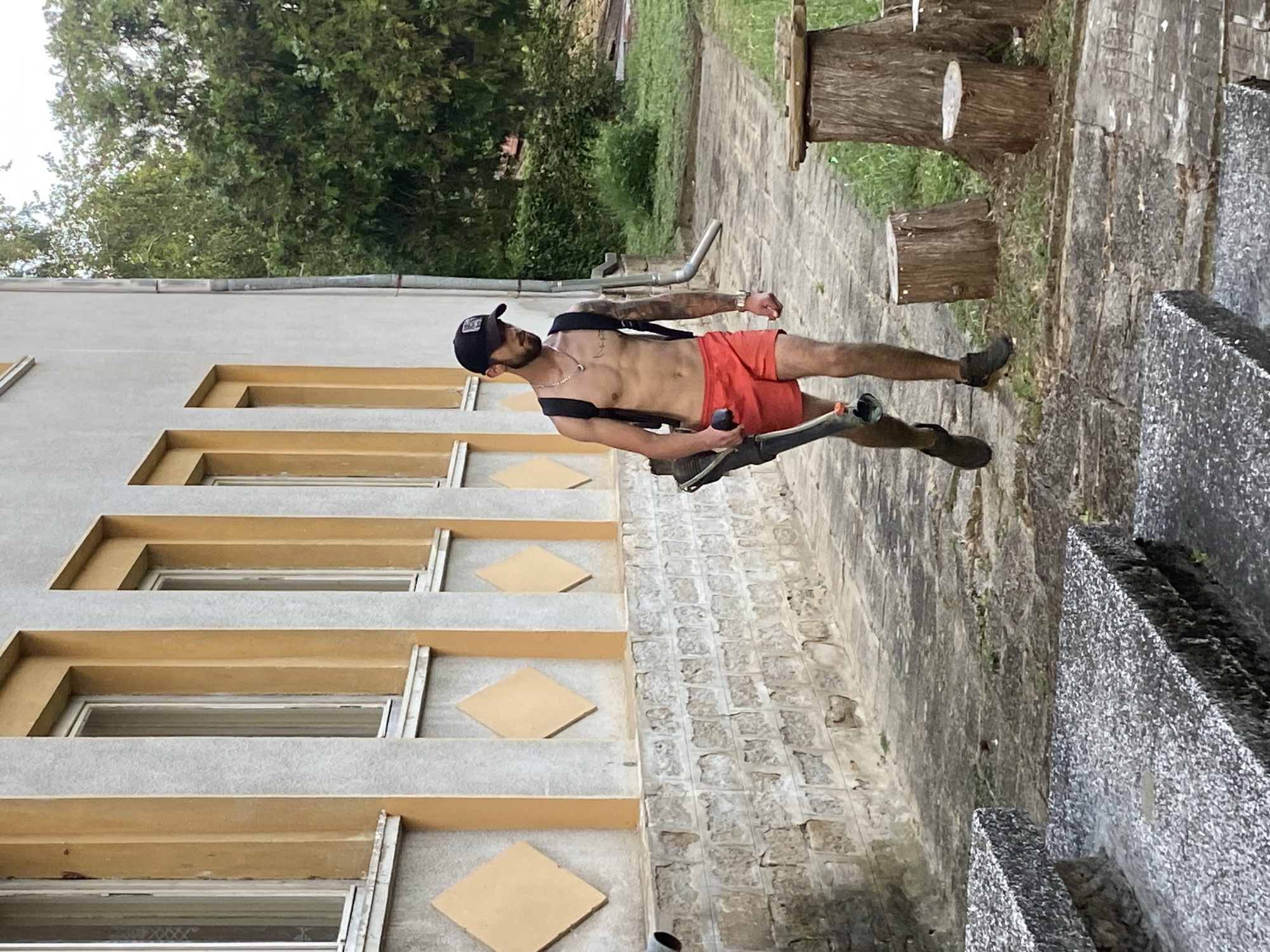 In Hirevo, Bulgaria, a shirtless young man with dark hair and a beard walks on a stone pathway next to a building with light gray and orange accents. He is wearing a black baseball cap, red shorts, and dark shoes, and carries a black backpack and a leaf blower. The pathway is bordered by a stone wall on the left and several tree stumps on the right. Green trees and foliage are visible in the background. The scene appears to be an outdoor setting, possibly a park or yard, adjacent to a building.