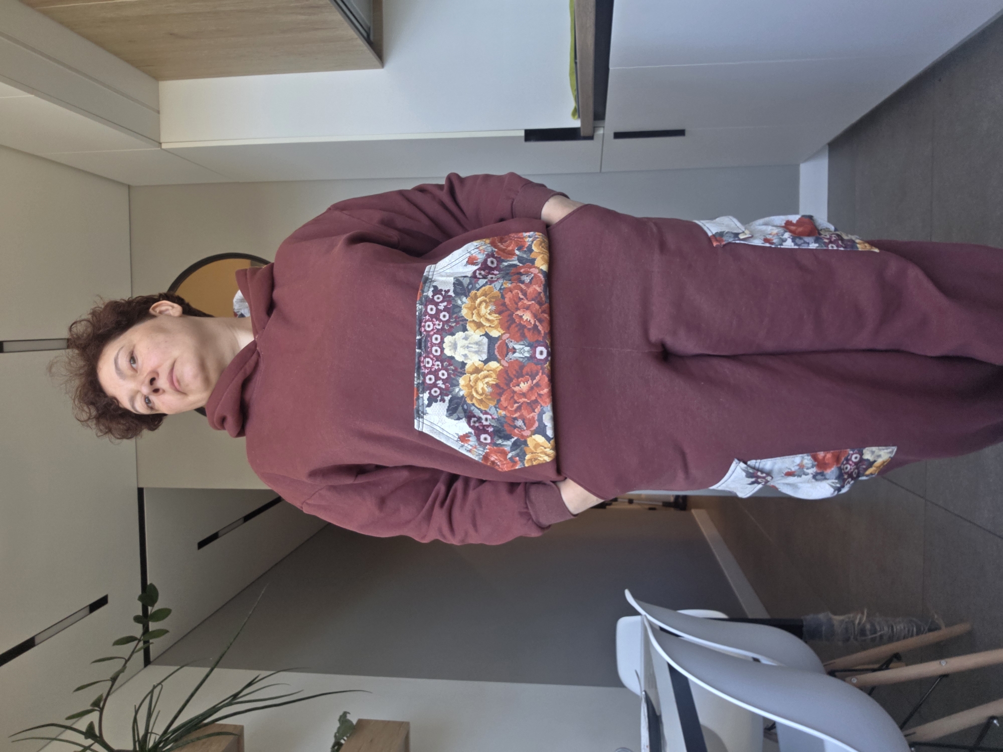 A person with short, curly brown hair, wearing a maroon hooded sweatshirt and maroon sweatpants with floral print panels on the sides, stands indoors. The location appears to be a modern apartment or house. Behind the person are white kitchen cabinets, a round mirror, a potted plant, and a gray wall. A white and gray chair is also visible. The image does not provide enough information to definitively determine if this is in Saint Petersburg, Russia.