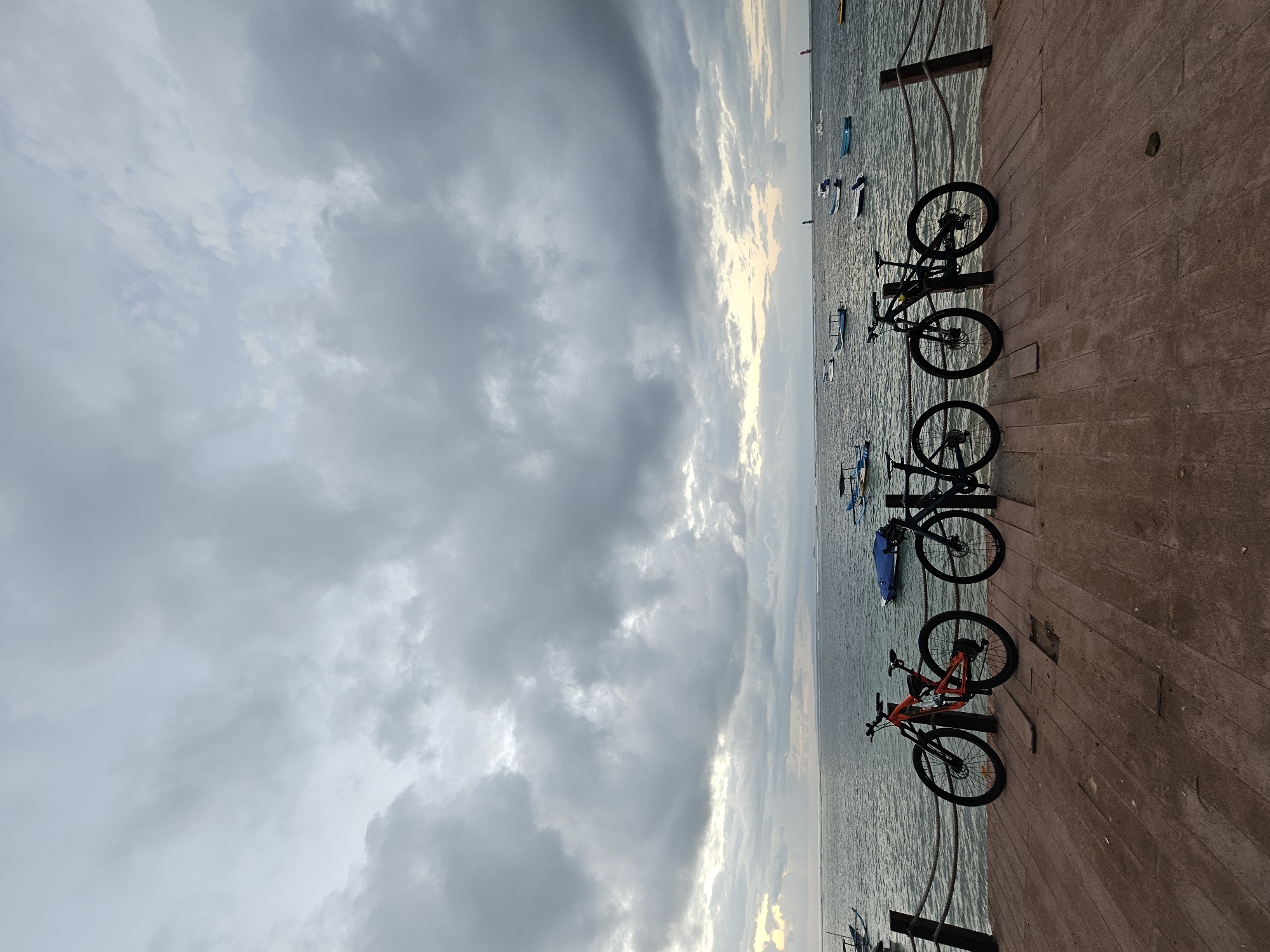 Three bicycles are parked on a wooden pier next to the ocean under a cloudy sky. The bicycles are all dark-colored mountain bikes, with one in the center being orange and black. The pier is made of wooden planks and is lined with a rope railing. In the background, the ocean is calm with several small boats floating on the water. The sky is overcast with heavy gray clouds, but there are hints of light breaking through on the horizon. The overall atmosphere is peaceful and serene. This scene is likely located on the coast of Denpasar, Indonesia, possibly at a beachside park or a harbor.
