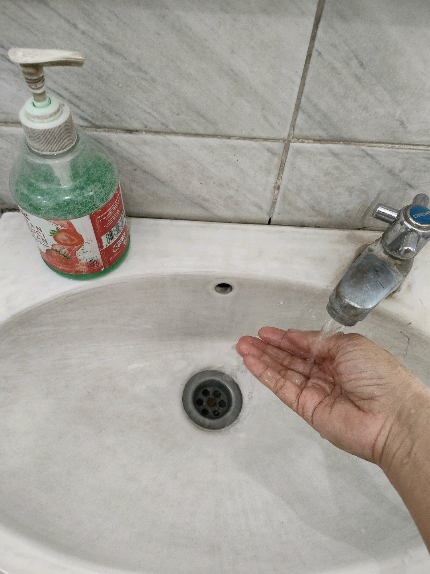 A close-up, high-angle shot shows a white ceramic sink in a bathroom. A hand is being washed under a stream of water from a chrome faucet. To the left of the sink is a clear plastic bottle of green liquid soap with a white pump dispenser. The background is a wall tiled with light gray and white marble. The overall scene is clean and well-lit, suggesting a typical Indonesian bathroom environment. The sink has a drain in the center.