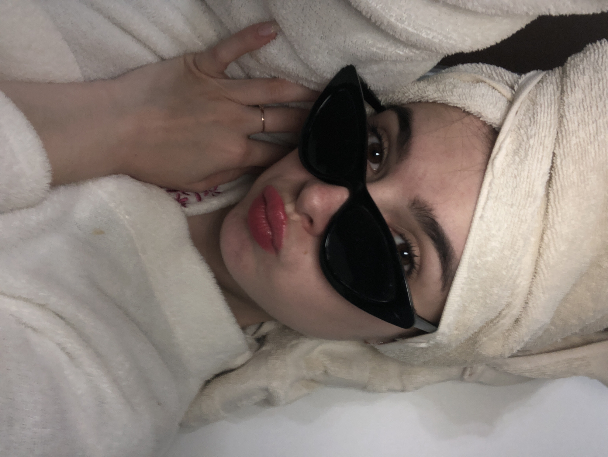 A woman with brown eyes and dark eyebrows is wearing red lipstick and black sunglasses. She is wrapped in a white towel on her head and wearing a white robe. She has a ring on her finger. The location is not discernible from the image.