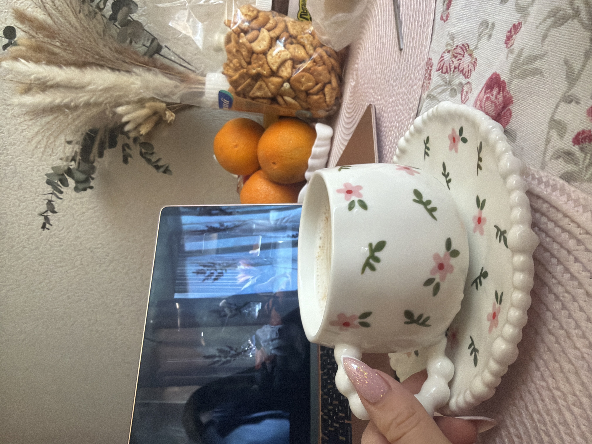 A cluttered scene features a laptop partially visible on the lower left, reflecting a window scene suggestive of Saint Petersburg, Russia, given its architectural style visible in the reflection. Adjacent to the laptop are stacked oranges and a bag of animal crackers. In the foreground, a hand with a decorated fingernail holds a white teacup with a floral pattern on a matching saucer. The backdrop includes dried floral arrangements and a patterned tablecloth.
