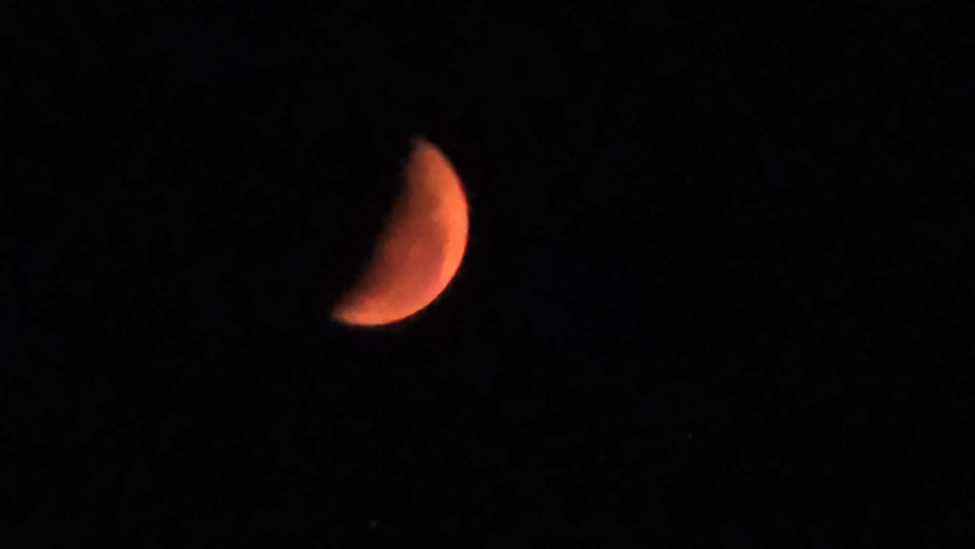 The image shows a crescent moon, reddish-orange in color, set against a dark black night sky. The moon is located towards the left side of the frame. Tiny, faint dots, possibly stars, are visible in the lower portion of the image. It is hard to determine the exact location of the city of Nha Trang, Vietnam.