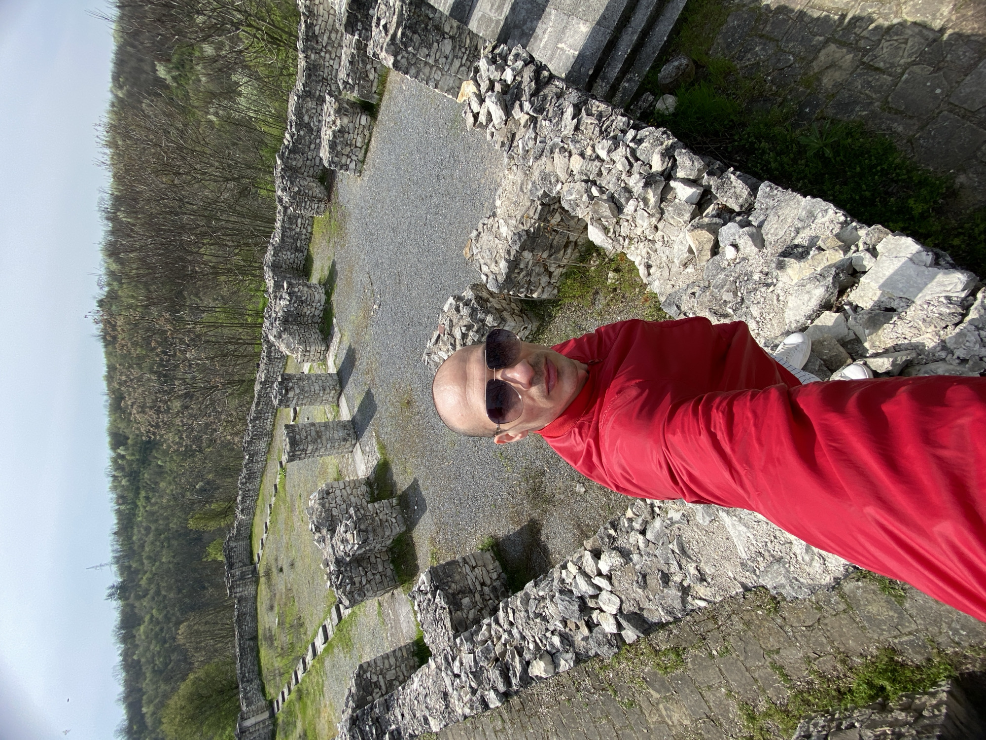 A man is posing for a photograph at the ruins of Storgozia, a Roman fortress located near the city of Pleven, Bulgaria. He is wearing a red shirt and sunglasses. The ruins consist of stone walls and staircases. The ground within the walls is covered in gravel and grass. A forest is visible in the background. The photo is taken from a high angle.