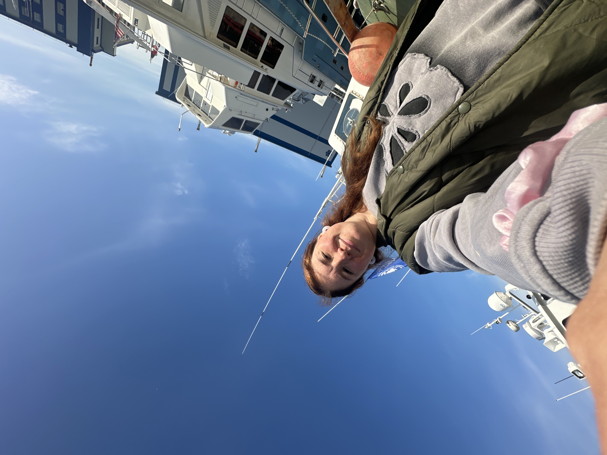 A woman with auburn hair, wearing a green puffer vest over a grey sweatshirt, is taking a selfie on a bright, sunny day. She is standing on a dock next to several boats. The boats are white with blue accents and appear to be docked in a harbor. The sky is a clear, vibrant blue with a few wispy clouds. There are tall, white antenna-like structures rising from the boats. In the background, industrial buildings can be seen. The overall setting suggests a maritime or industrial area within the city of Petropavlovsk-Kamchatsky, Russia.