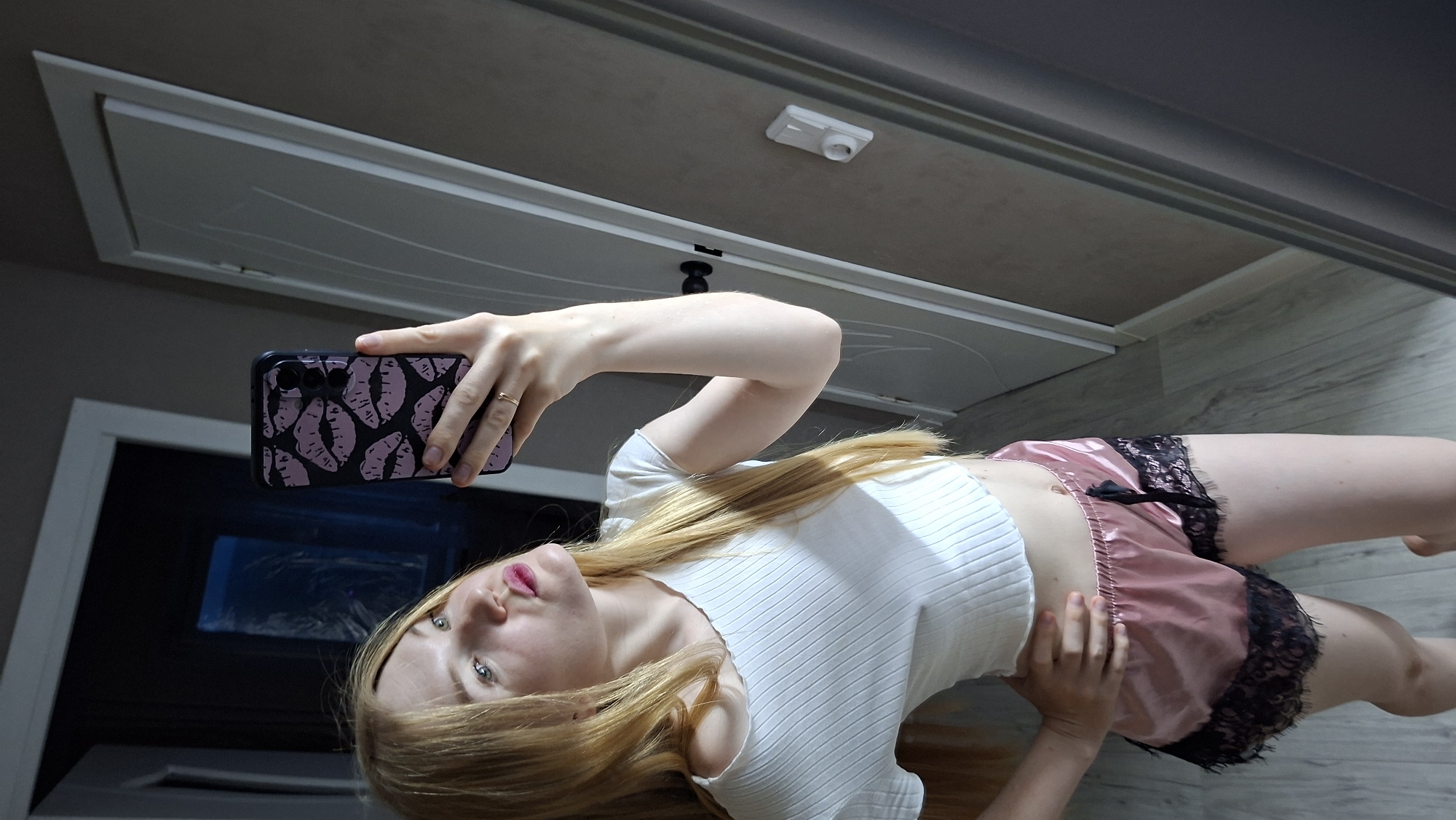 In this media file, a young Caucasian woman with long blonde hair and blue eyes is taking a mirror selfie. She is wearing a white ribbed crop top and pink satin shorts with black lace trim. Her hair is falling over her shoulders, and she is looking directly at the camera with a slight pout. She is holding a smartphone in her right hand, which has a black case with a pink lip pattern. A thin gold ring is visible on her left hand.
The background appears to be an indoor setting, possibly a bedroom or hallway, with light-colored wooden floorboards. A closed door with white paneling is visible behind her. The lighting suggests it could be daytime. The overall mood is casual and playful. There is no visible text in the image.