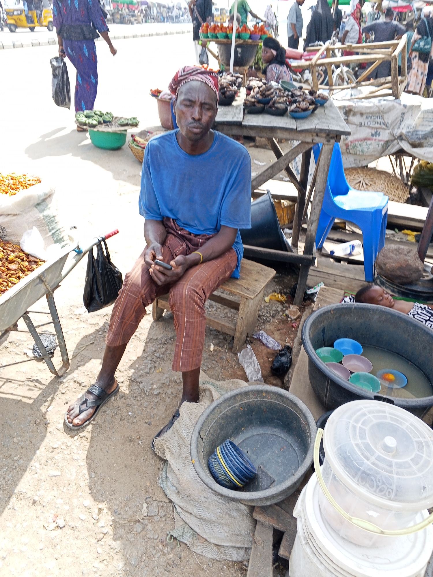 The image captures a market scene in Zango-ndaji, Nigeria. A man wearing a blue t-shirt and brown plaid pants sits on a wooden stool, holding a smartphone. He has a red and white head covering. To his left is a wheelbarrow with what appears to be foodstuff in a white bag. To his right are stacked buckets and tubs; one tub contains colorful bowls submerged in water, and a baby sleeps in the background near the tub. Behind the man is a table displaying bowls of dark food items and a blue plastic chair. In the background, other market vendors and shoppers are visible.