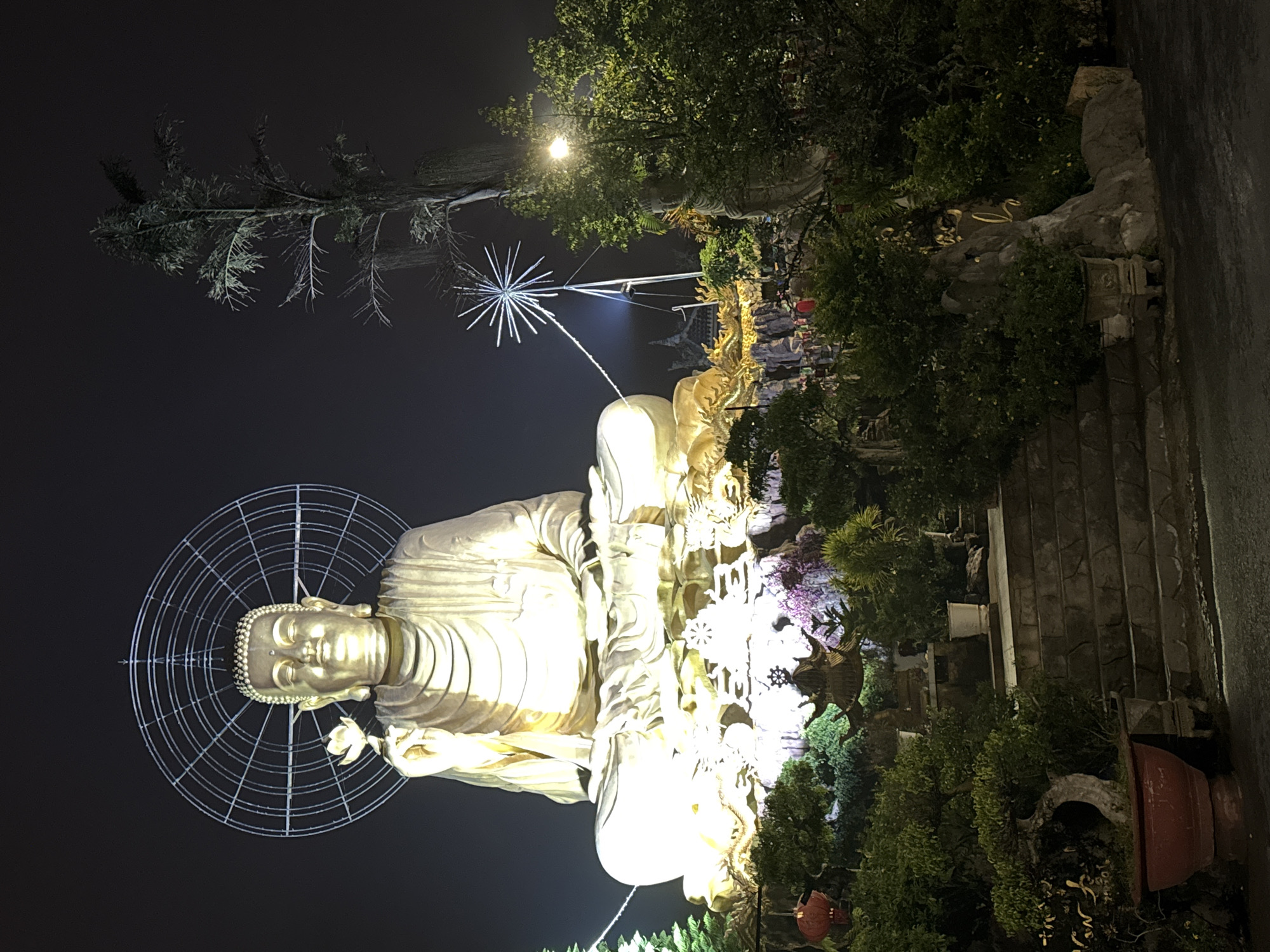 This nighttime image, taken in Lâm Viên - Đà Lạt, Vietnam, features a monumental golden Buddha statue as its central subject. The Buddha is depicted in a seated, meditative posture, holding a lotus flower, and is brilliantly illuminated, glowing against the dark sky. A striking circular grid, appearing to be made of light or metal, forms a halo behind the Buddha's head.
The statue is situated outdoors within a lush, landscaped environment, surrounded by dense trees and bushes. To the right of the Buddha, a tall pole supports a prominent, modern-looking starburst light fixture, adding to the decorative lighting of the scene. The base of the Buddha statue appears to be adorned with intricate golden carvings or smaller statues. In the foreground to the right, a stone pathway or steps are visible, bordered by greenery and a large red planter pot. The overall setting suggests a temple or a beautifully illuminated garden at night, creating a serene and awe-inspiring atmosphere. There is no visible text in the image.