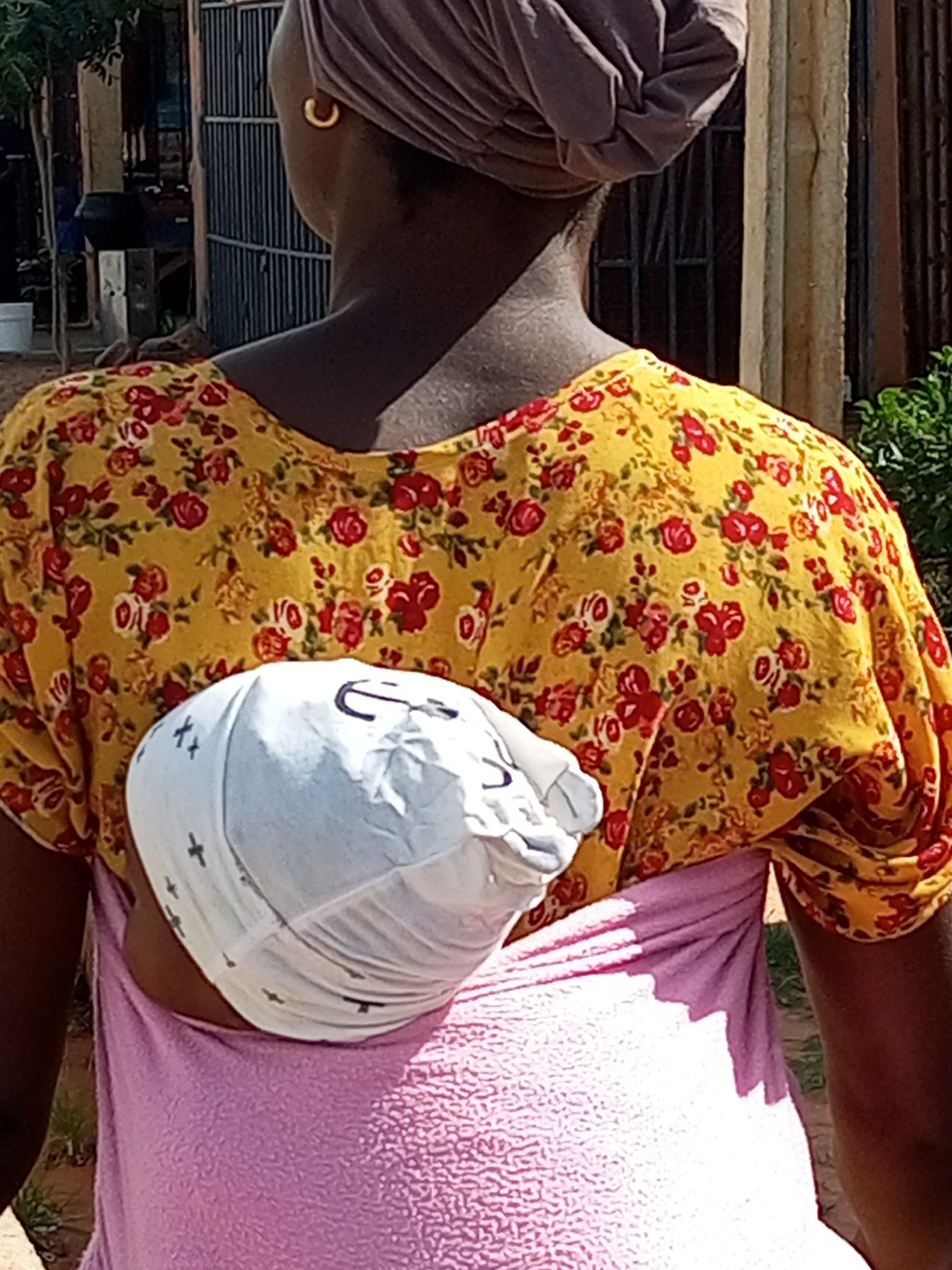 The image shows a close-up of a woman in Tempure, Nigeria, carrying a baby on her back. The woman is wearing a yellow floral print shirt and a pink fabric wrap. The baby is wrapped in a white cloth. The background appears to be a street scene with a fence and some greenery.