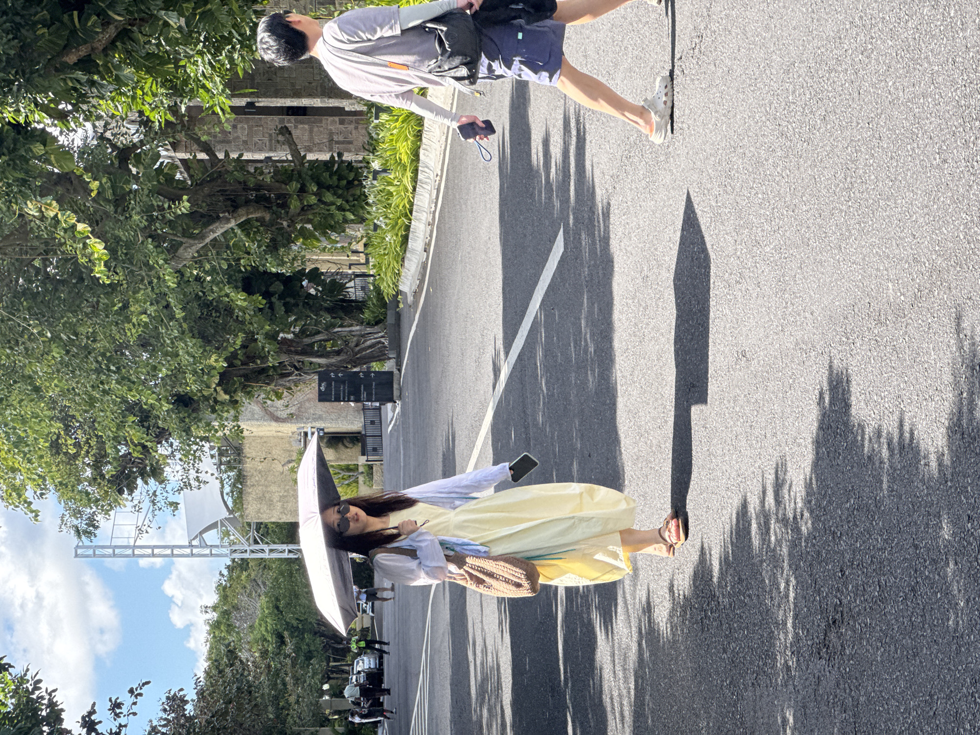 The image depicts a street scene in Kuta Selatan, Indonesia, on a sunny day. In the foreground, a woman walks towards the viewer, holding a partially open umbrella over her head. She is wearing a yellow dress, a light blue cardigan, sunglasses, and sandals. A woven bag is slung over her shoulder, and she holds a smartphone in her right hand. Behind her and to the right, a man walks away from the viewer. He is wearing a striped t-shirt, blue shorts, and white Crocs. He carries a dark bag on his shoulder and holds a smartphone in his left hand. The street is paved with asphalt and has white lane markings. Lush green trees and vegetation line the sides of the road, with a stone building visible in the background. Other people and vehicles are present further down the street. The sunlight casts distinct shadows on the asphalt, indicating a bright and clear day.