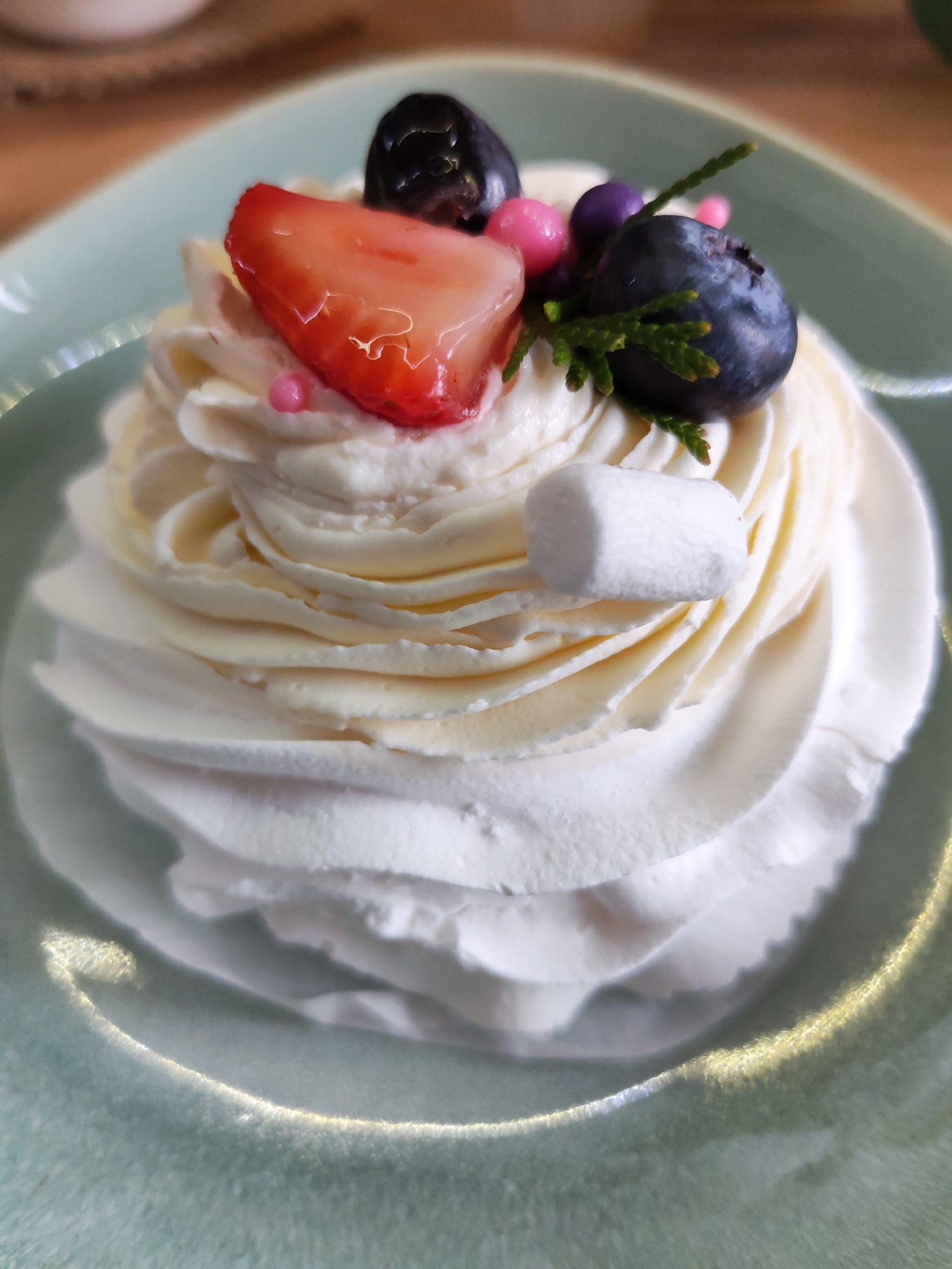 The image shows a dessert sitting on a pale green plate. The dessert is composed of white swirled cream, and is topped with a slice of strawberry, two blueberries, a sprig of green leaves, pink and purple sprinkles, and a single marshmallow. The photograph was not necessarily taken in the city of Izhevsk, Russia.