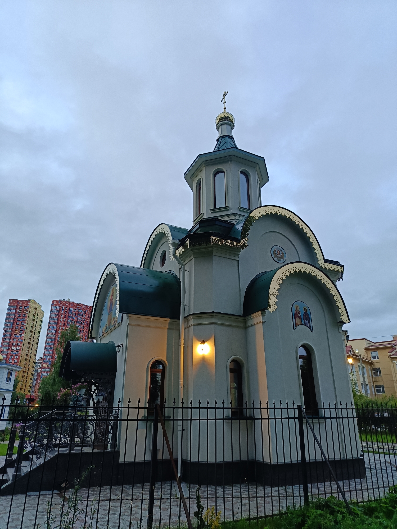 The image depicts the Church of the Icon of the Mother of God "Kazanskaya" located at 35 Leninsky Prospekt in Saint Petersburg, Russia. The church, viewed from a low angle, features a light gray facade, a dark green roof, and a golden dome topped with a cross. Decorative elements with golden trim are visible along the rooflines. Adjacent to the church, two colorful, modern high-rise buildings stand, one yellow and red, the other red and yellow. The scene is set against a cloudy, overcast sky. The church is enclosed by a black metal fence, with a paved walkway visible behind it. The lighting suggests it is dusk or an overcast day. No people are visible in the image.
