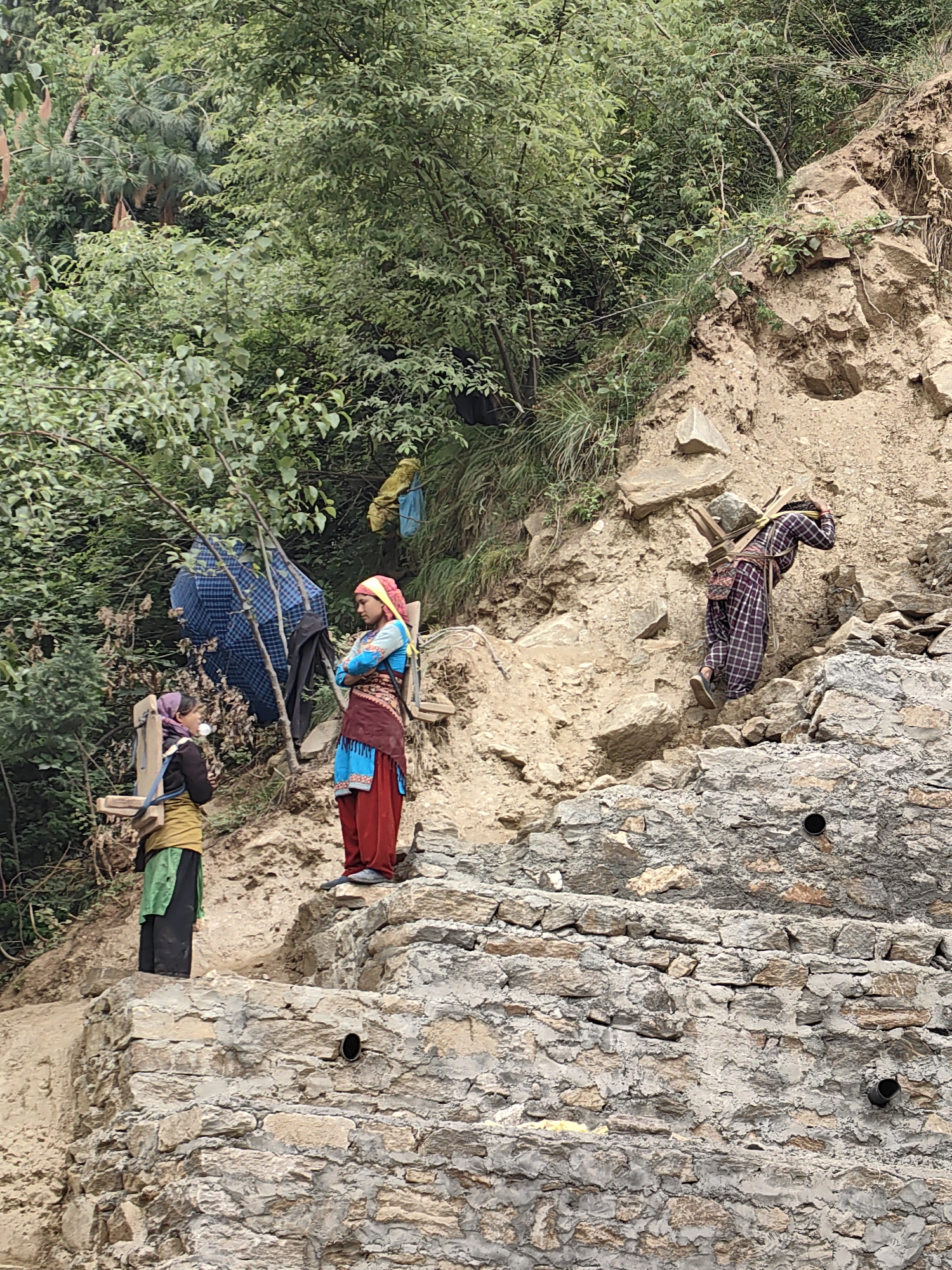 In Vashisht, India, three women are working on a rocky, inclined slope. The woman on the far right, wearing a plaid dress and headscarf, carries a wooden frame loaded with rocks on her back, ascending the slope. In the middle ground, a woman in a red skirt and blue top with a pink headscarf stands with her arms crossed, facing left. To her left, another woman in a yellow top and green skirt with a wooden contraption on her back faces forward, looking down. A blue and black checkered umbrella is propped up between the two standing women. The environment is rugged, with loose soil, rocks, and vegetation. The background consists of dense green foliage and trees.