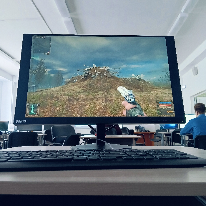 The image displays a computer workstation located within a classroom or computer lab in Chaikovsky, Russia. In the immediate foreground, a black computer keyboard rests on a light-colored desk. Behind the keyboard, a black ThinkVision monitor occupies the central view, displaying a first-person shooter video game. The game scene depicts a player's gloved hand holding a white and silver handgun, aimed at a desolate, brown, hilly landscape featuring a large mound topped with a structure of metal debris and antennas under a heavily clouded sky. Visible in-game interface elements include a mini-map in the upper left, a character status icon in the lower left, and ammunition count "2/882" in the lower right.
In the background, on the right side of the frame, one individual is seated at another computer station, facing away from the camera. This person is wearing a blue long-sleeved shirt. Additional computer setups with black office chairs are partially visible further back in the room, which features white walls, white ceiling tiles with fluorescent lighting fixtures, and a white-framed doorway on the far right.