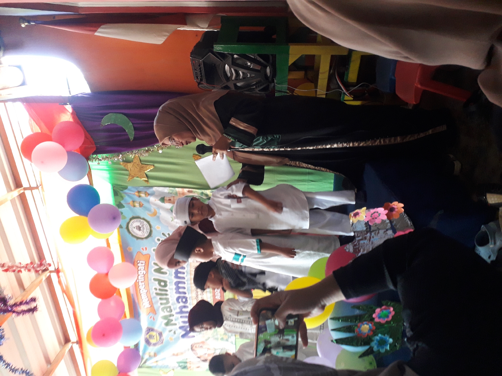 An outdoor event is taking place under a wooden canopy structure in Cankuang, Indonesia. The event appears to be a celebration for Maulid Muhammad, as indicated by a banner that reads "Memperingati Maulid Muhammad". A group of children in white traditional clothing are lined up, likely for a performance or presentation. A woman in a hijab is speaking into a microphone, holding a paper, suggesting she is a speaker or presenter. Balloons in various colors adorn the background, and decorations like colorful flowers and a crescent moon are visible. In the foreground, a person is recording the event on a smartphone. The overall atmosphere suggests a community gathering, possibly a school or religious event.