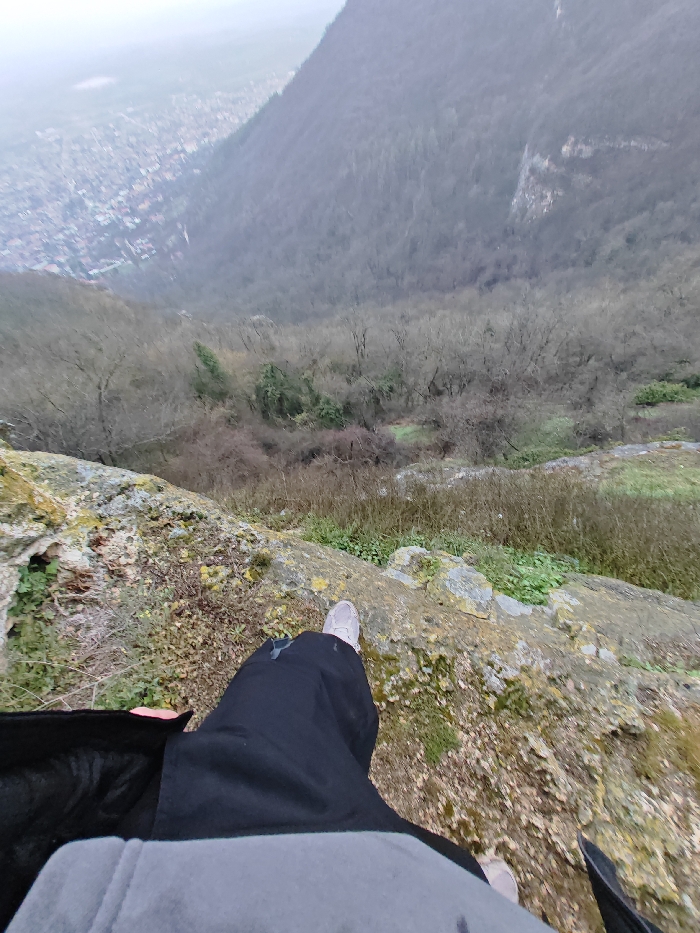 The image presents a high-angle, first-person perspective from a rocky cliff edge, overlooking a valley containing the city of Shumen, Bulgaria, and adjacent forested terrain. In the immediate foreground, the lower torso of an individual, dressed in a grey top and black pants, is visible. A light-colored athletic shoe is present on the left foot, positioned on a rugged, moss-covered rock formation. The ground immediately beyond the ledge drops sharply into a steep, densely forested slope, characterized predominantly by bare deciduous trees with some scattered evergreen foliage. To the left in the background, the urban area of Shumen spreads across the valley floor, appearing somewhat indistinct due to atmospheric haze. To the right, a large, tree-covered mountain or plateau side rises, featuring a prominent, lighter-colored rocky outcrop on its face. The sky is uniformly overcast, contributing to subdued lighting conditions.