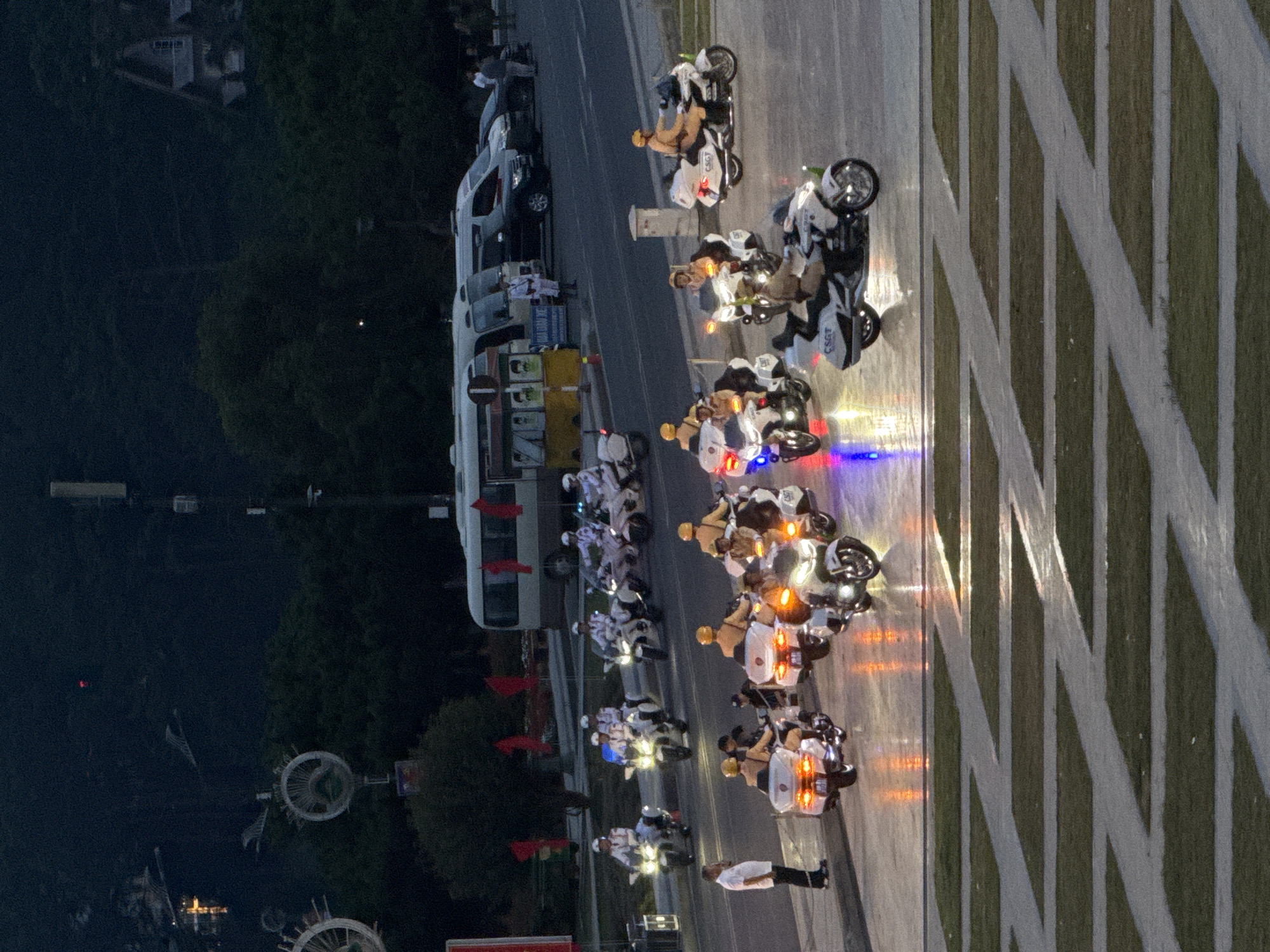 In Da Lat, Vietnam, at dusk, a formation of police motorcyclists in white uniforms with white helmets are captured on a paved area with illuminated square grass patches. The group is riding a line of white police motorcycles, several of which have flashing blue and red lights. Another group of police motorcyclists in tan uniforms and tan helmets are also visible, riding similar motorcycles. One individual in a white shirt and black pants stands on the left, observing the motorcyclists. In the background, several buses and cars are parked on a road, and trees and buildings are visible on a hillside under a dark sky.
