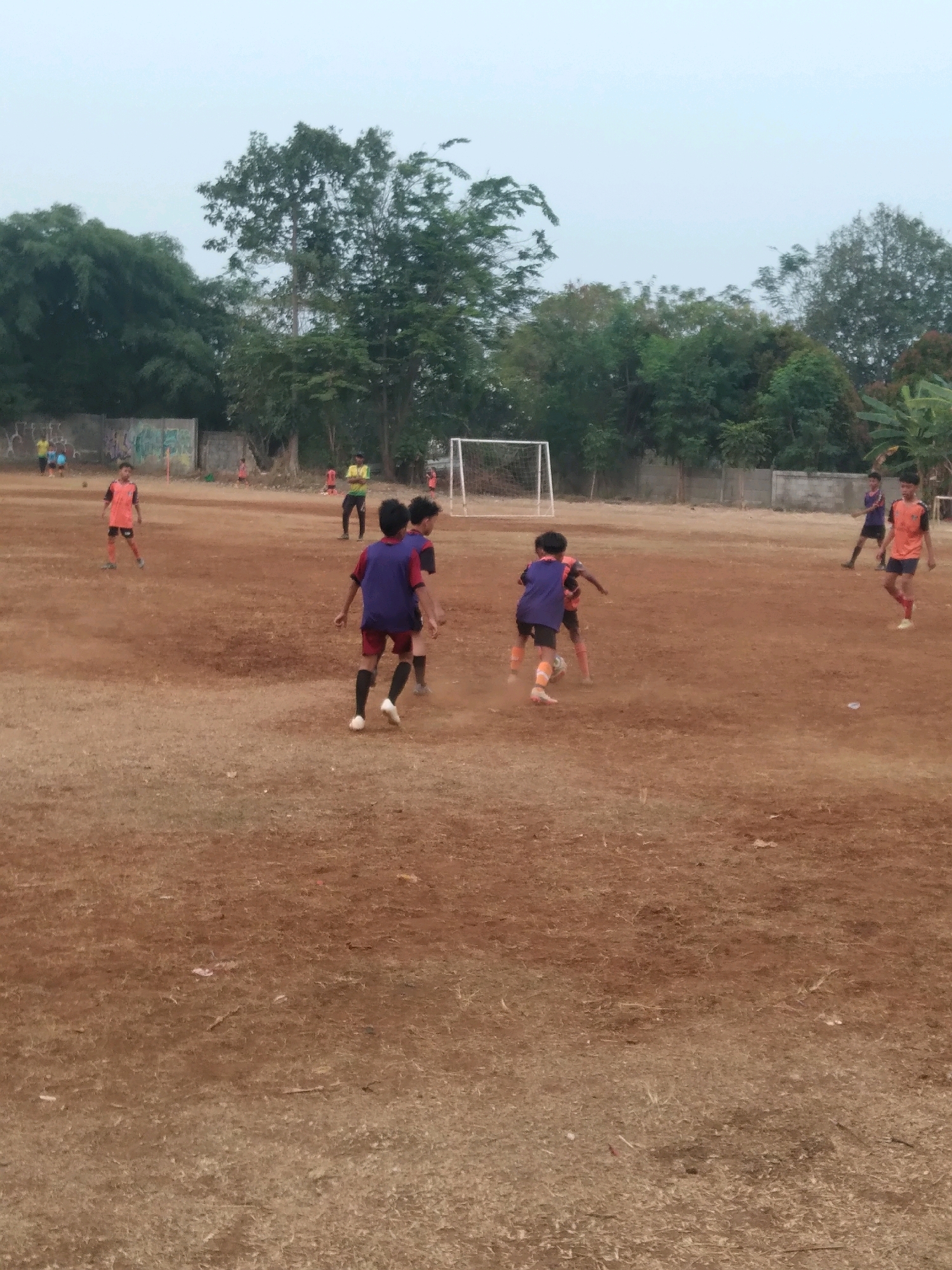 This image depicts a group of young boys playing soccer on a dirt field. The field appears to be an informal sports ground located in South Tangerang, Indonesia. Several players are visible, most wearing orange and blue jerseys. One boy in the foreground is wearing a blue jersey and red shorts, actively kicking a soccer ball. Another boy in an orange jersey and black shorts is running nearby. In the background, more players are scattered across the field, some walking and others running. A soccer goal is visible in the distance. The environment is characterized by a dirt field with sparse dry grass, surrounded by trees and a fence. The sky is overcast, suggesting it may be early morning or late afternoon. The overall scene captures a moment of casual soccer play in a local setting in South Tangerang.