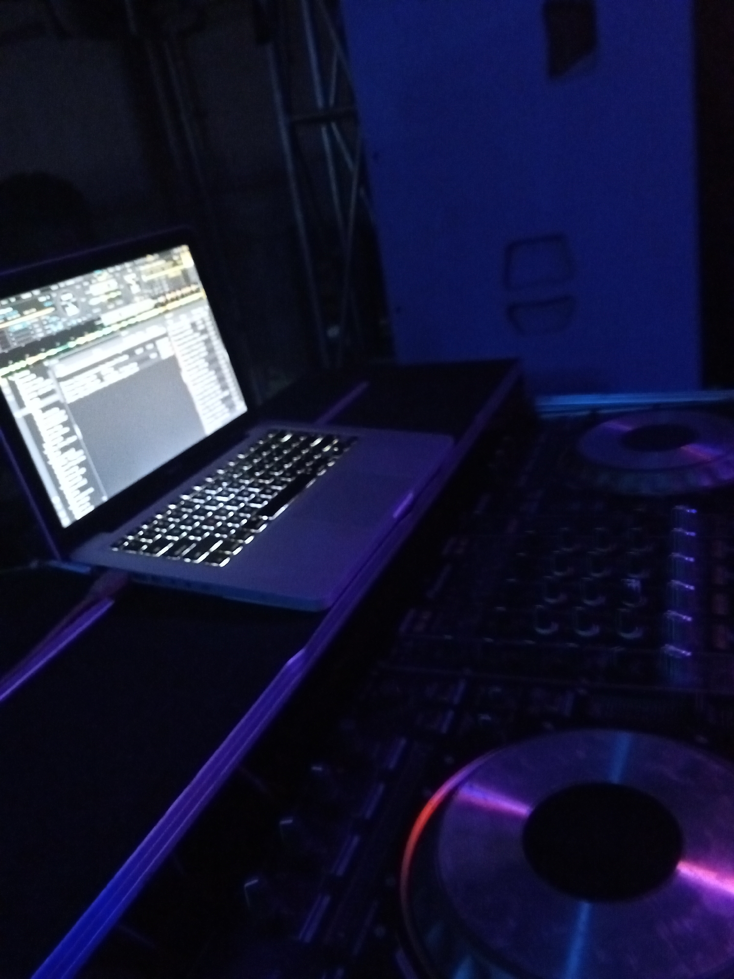 A laptop with its screen displaying audio mixing software rests on a case next to a DJ controller. The scene is illuminated by blue and purple lighting. There is no evidence to suggest this is located in Lokoja, Nigeria.