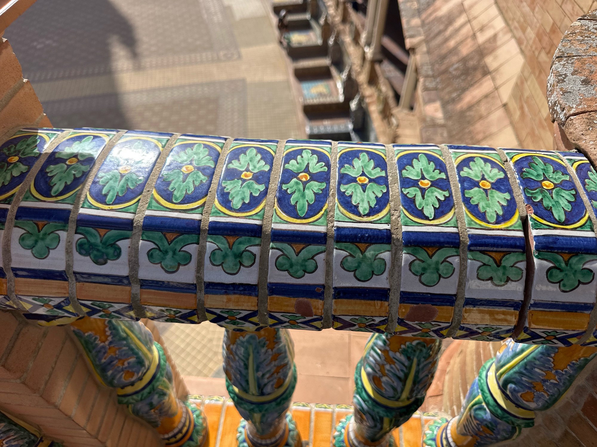 The image shows a close-up of a decorative ceramic railing on a staircase in Seville, Spain. The railing is adorned with hand-painted tiles featuring a repeating pattern of green floral motifs with blue borders and yellow accents. The tiles are set against a brick structure. Below the railing, the staircase is supported by elaborately decorated ceramic balusters, also painted in vibrant blues, greens, and yellows, with patterns reminiscent of foliage. The background reveals the upper part of a staircase with patterned flooring and brick walls, suggesting an architectural setting within the city. The sunlight casts shadows on the tiled surface, indicating an outdoor environment.