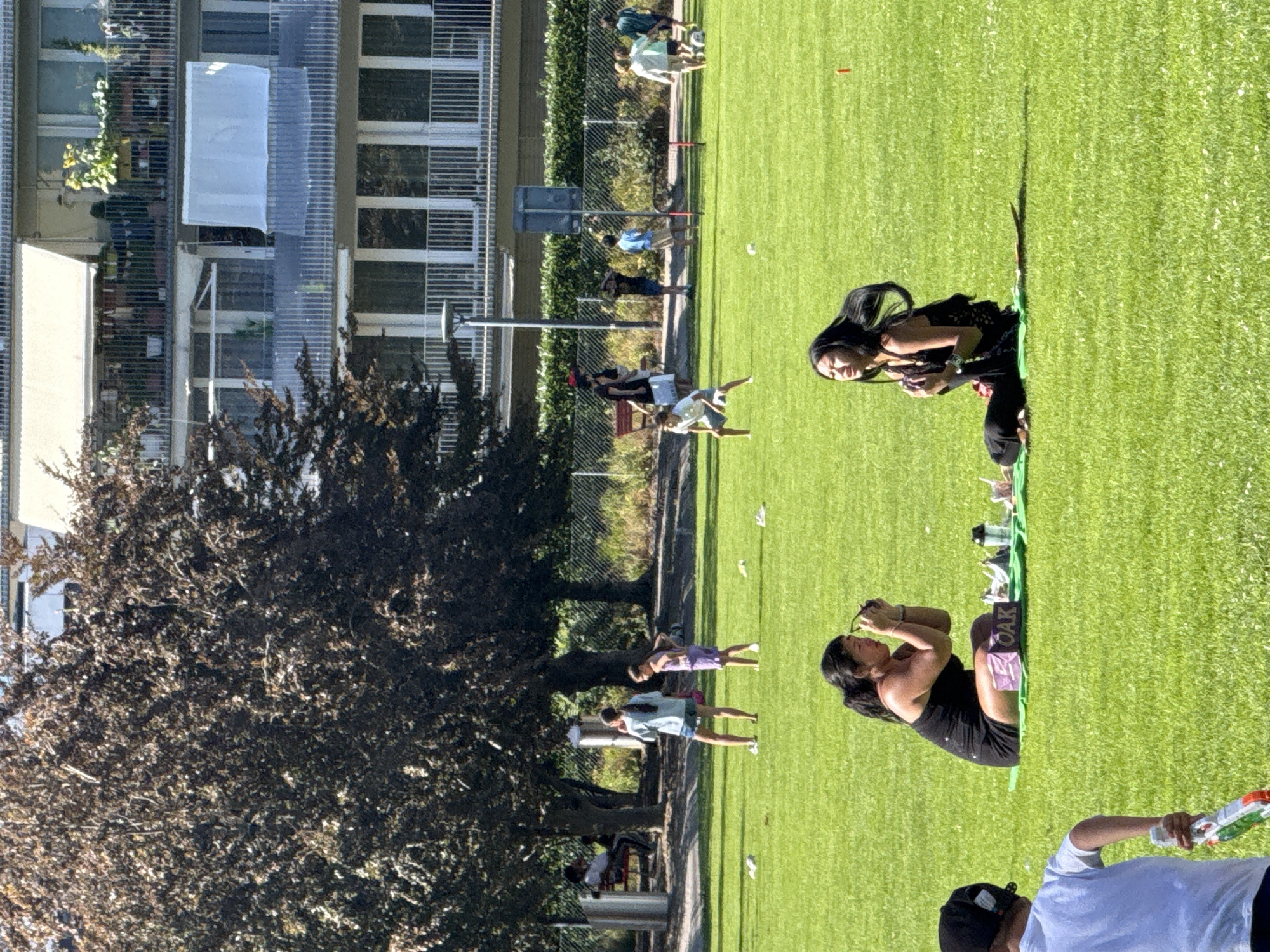 The image depicts an outdoor scene on a sunny day at a park in Zurich, Switzerland. In the foreground, two women are seated on a green lawn. One woman, wearing a black dress, is adjusting her sunglasses. Next to her is a small purple box with the word "OAK" printed on it, and some items on a green mat. The other woman, wearing a black top and pants, is meditating with her eyes closed and hair blowing in the wind. A young person wearing a white t-shirt and a black baseball cap is visible from the back on the left, holding a toy.
In the middle ground, several children and adults are scattered across the park. Some children are playing with a soccer ball, while others are standing and interacting. There are trees with dark foliage and a fence in the background.
Behind the park, a multi-story building with balconies is visible. The balconies are adorned with plants and awnings, suggesting a residential area. The overall impression is a relaxed atmosphere of people enjoying a day outdoors in a park.