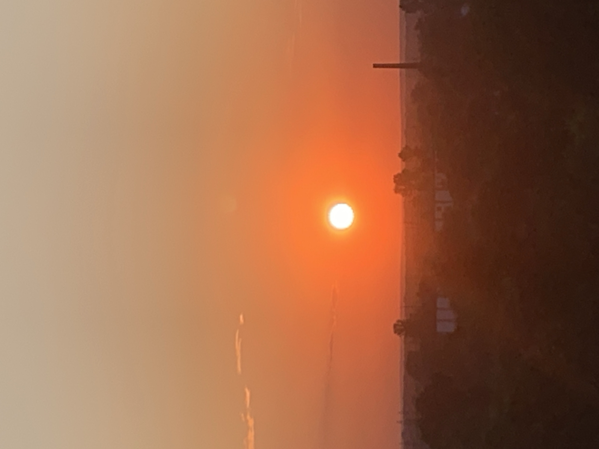 The image shows a sunset over a city, likely Kolomyia, Ukraine, with the sun positioned near the center of the frame. The sky is a gradient of warm orange and peach tones, with a few faint clouds visible. Below the sky, a hazy silhouette of buildings and trees is visible, with a tall, slender chimney or tower standing out on the right side of the image. The overall scene is serene and atmospheric.