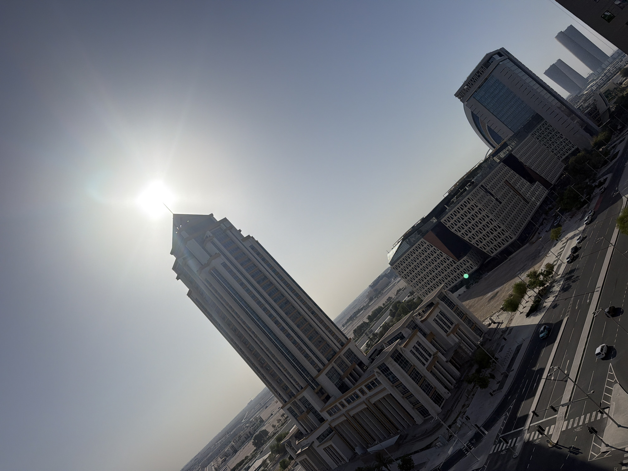 This elevated, diagonally oriented image captures a bustling urban landscape under bright daytime conditions in the city of Unknown, Unknown. The scene is dominated by modern architecture and infrastructure, bathed in direct sunlight from the upper right, creating a distinct lens flare.
The sky, occupying the upper portion of the frame, transitions from a pale blue near the horizon to a lighter, almost white hue around the intensely bright sun. No clouds are visible.
A prominent, tall skyscraper, featuring a light-colored facade with numerous vertical rows of rectangular windows and a distinctive pointed, multi-tiered apex, rises diagonally across the left-center of the image. Surrounding its base are several interconnected mid-rise buildings, constructed in a similar architectural style, forming a complex of structures.
To the right of the tall skyscraper, in the mid-ground, a cluster of modern buildings includes one with a curved roofline and extensive glass paneling, and another with a patterned, grid-like facade. Further in the distance, several uniformly shaped, rectangular high-rise towers are visible, somewhat obscured by atmospheric haze.
At ground level, a wide, multi-lane road with clear white lane markings and pedestrian crosswalks stretches across the foreground. Traffic lights on poles are visible along the road. Several vehicles, including a white sedan, a dark SUV, and other cars, are present on the road, some appearing parked or in motion. Patches of green vegetation, including trees and bushes, are strategically placed along roadsides and between buildings. Beyond the immediate developed areas, expanses of arid, undeveloped land are visible, suggesting ongoing urban expansion. No individuals are distinctly visible in the image.