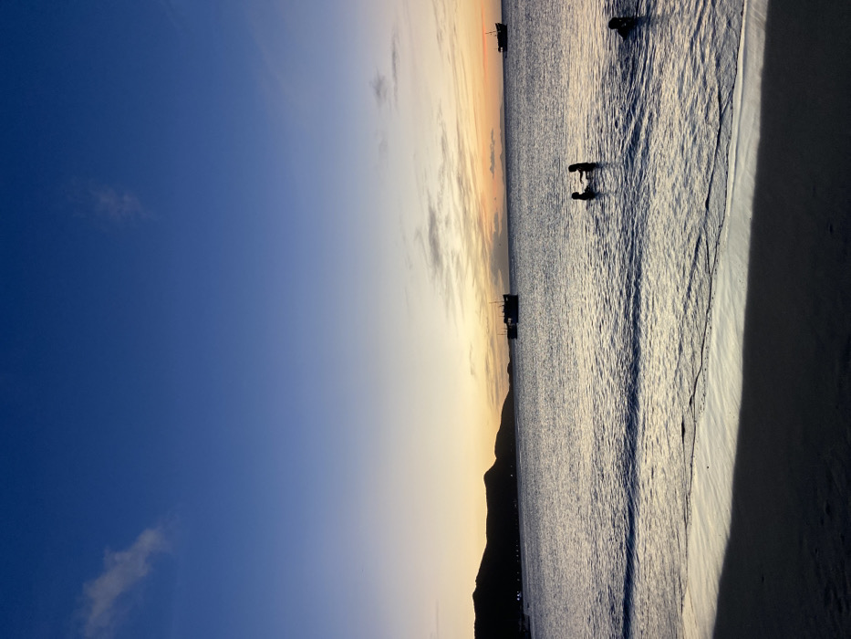 A serene beach scene unfolds during sunset. The sky transitions from deep blue to pale orange near the horizon. Silhouettes of distant mountains and a few boats are visible on the water. Two figures stand in the shallow water, facing each other. The beach itself is bathed in soft light, casting long shadows. Gentle waves create patterns on the sand.