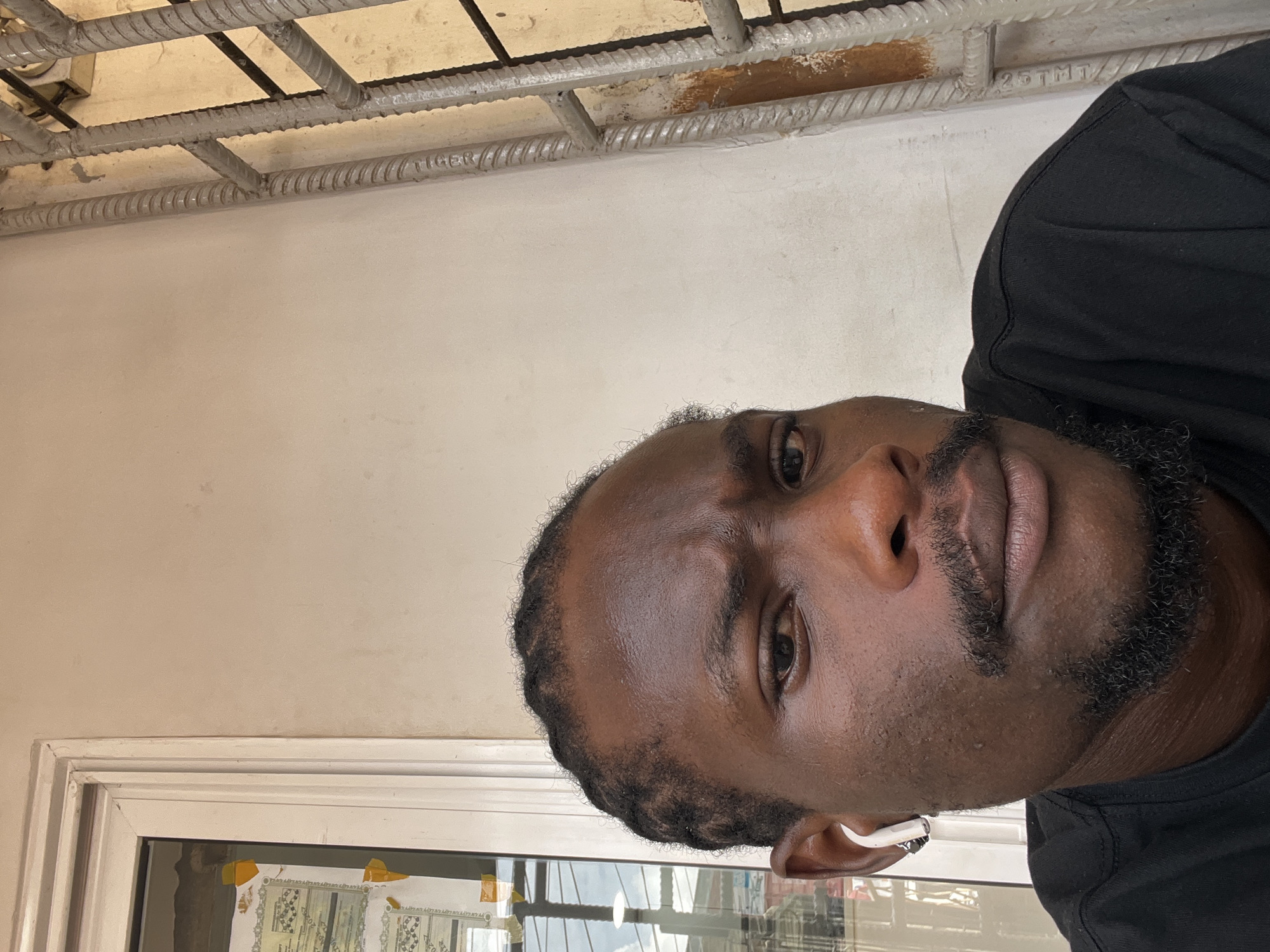 A medium, close-up shot shows a man with dark skin, short hair and a black beard, wearing a black shirt and a white earbud in his right ear. He is looking at the camera. Above him is a white ceiling with a grid of metal rebar. Behind the man to the left is a white wall, and below him is a window frame and a glimpse of the outside, potentially in Ikeja, Nigeria. The image is rotated slightly counter-clockwise.
