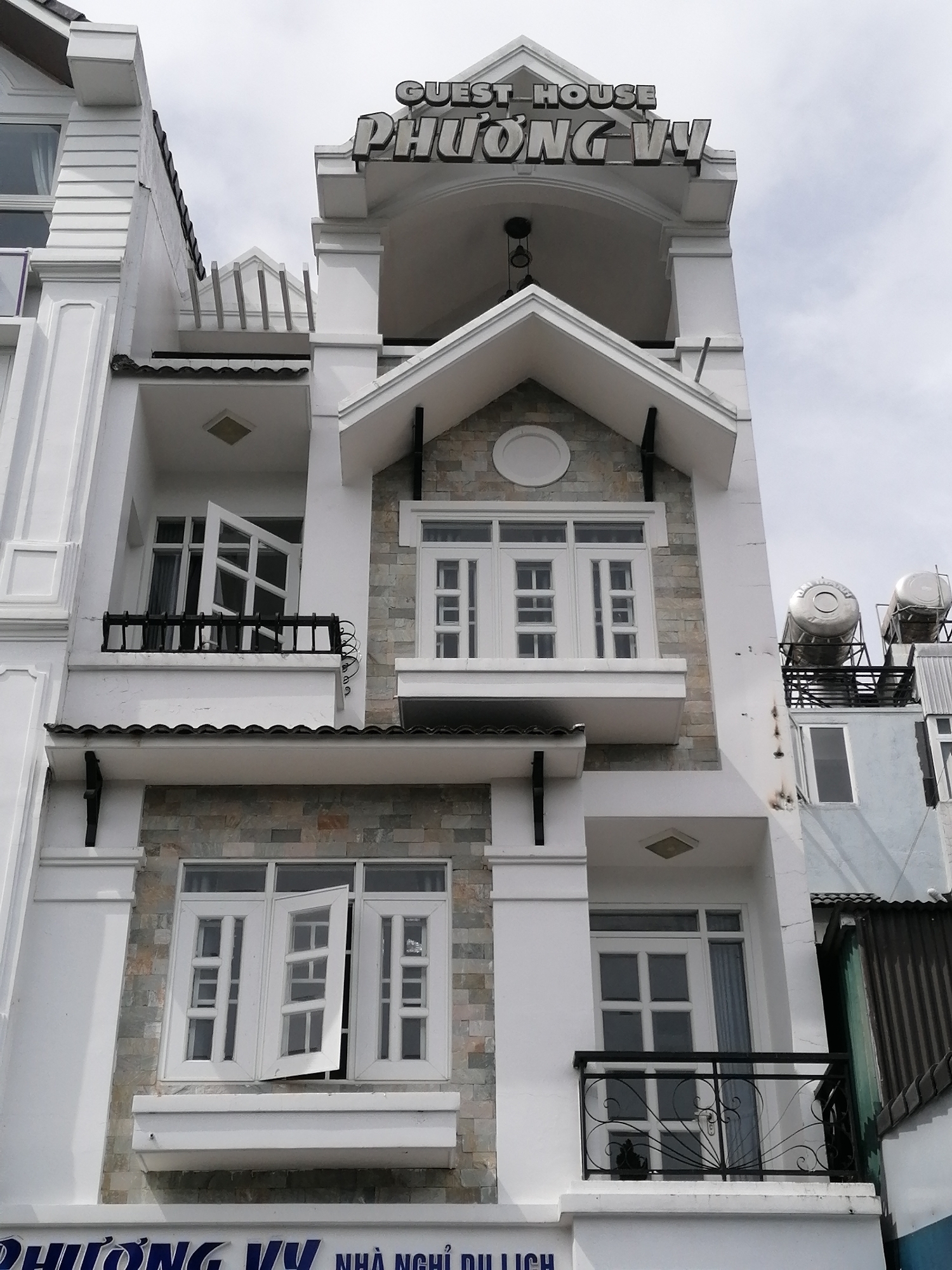 A multi-story guest house, identified by the sign "GUEST HOUSE PHUONG VY", is centrally featured against a cloudy sky. The building is predominantly white with stone accents on its facades. It has multiple balconies with decorative black iron railings. The windows are white-framed and appear to be made of multiple panes. On the rooftop, there are two large cylindrical water tanks. The architecture suggests it is located in an urban area, likely Da Lat, Vietnam, given the "PHUONG VY NHA NGHI DULICH" text at the bottom which translates to "Phuong Vy Tourist Guest House". No people are visible in the image. The overall impression is of a well-maintained, modern building.