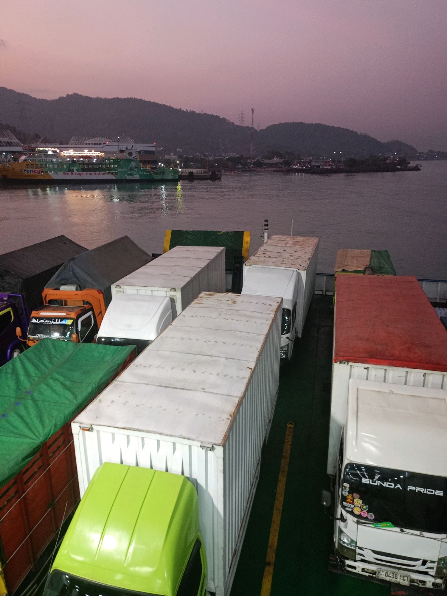 In the city of Cilegon, Indonesia, at dusk, a ferry is transporting numerous trucks and containers across a body of water. The sky exhibits a pale purple hue, indicative of twilight. The ferry's deck is densely packed with vehicles. Dominating the foreground are several large truck trailers, some covered with tarpaulins of various colors, including green and red, while others are white, revealing their corrugated metal construction. The cabs of the trucks are also visible, some with colorful decals and text. One white truck prominently displays "SUNDA PRIDE" on its windshield. In the background, a large passenger ferry with "ferizy" and "We Bridge the Nation" printed on its side is docked or approaching a port. The surrounding landscape features a hilly coastline with scattered lights and structures, suggesting a developed urban or industrial area. The water reflects the dim light from the sky and the ferry's lights, creating a muted sheen. The overall scene depicts a functional, logistical operation at the end of the day.
