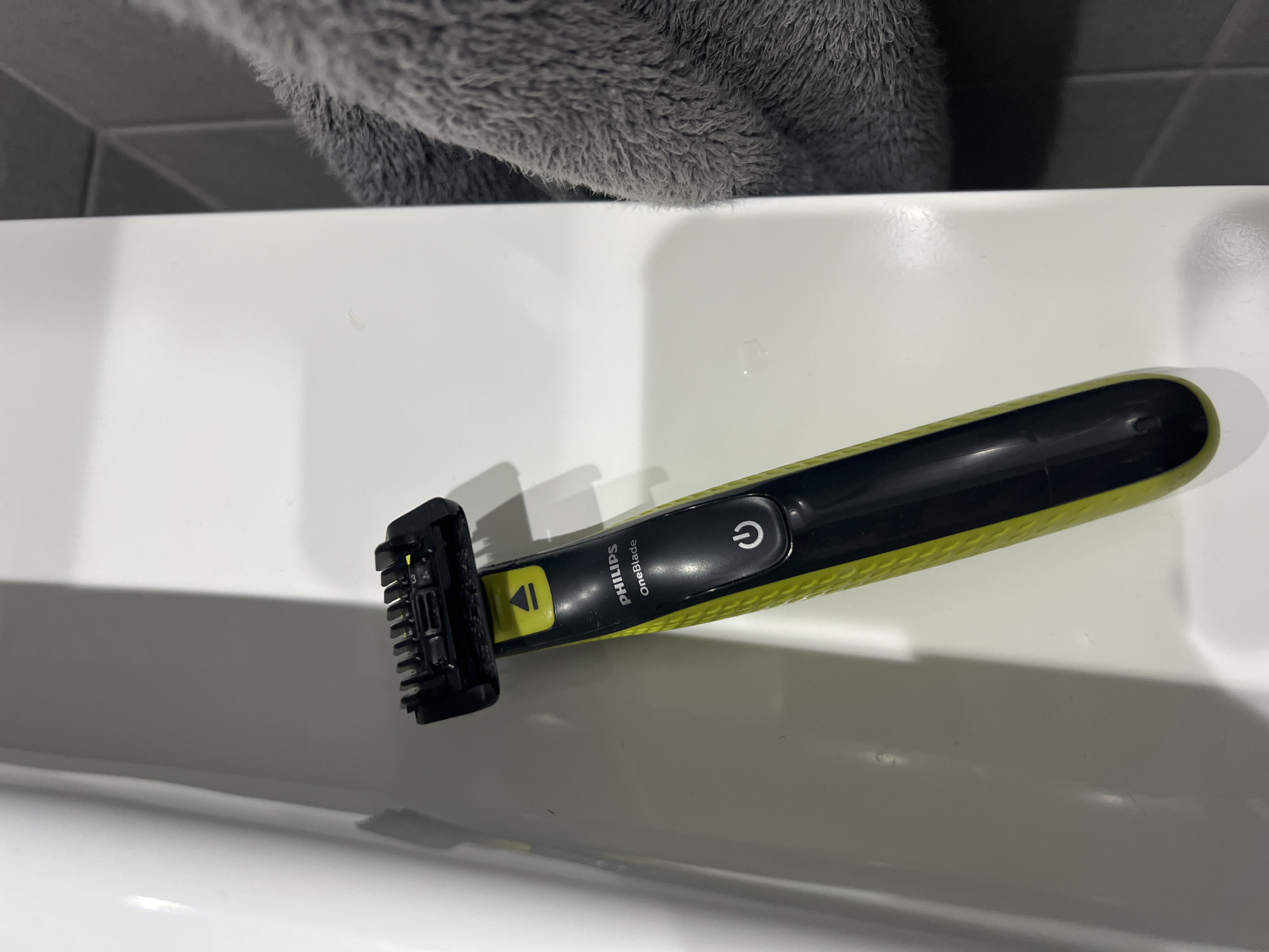 A Philips OneBlade electric razor sits on a white, glossy surface, possibly a bathroom sink or countertop. The razor is black and lime green with a black trimming attachment at one end. A gray, fluffy object, possibly a towel or fabric, is partially visible in the upper part of the frame. The background includes a glimpse of gray tiles. The location in Burgas, Bulgaria is not determinable from the image.