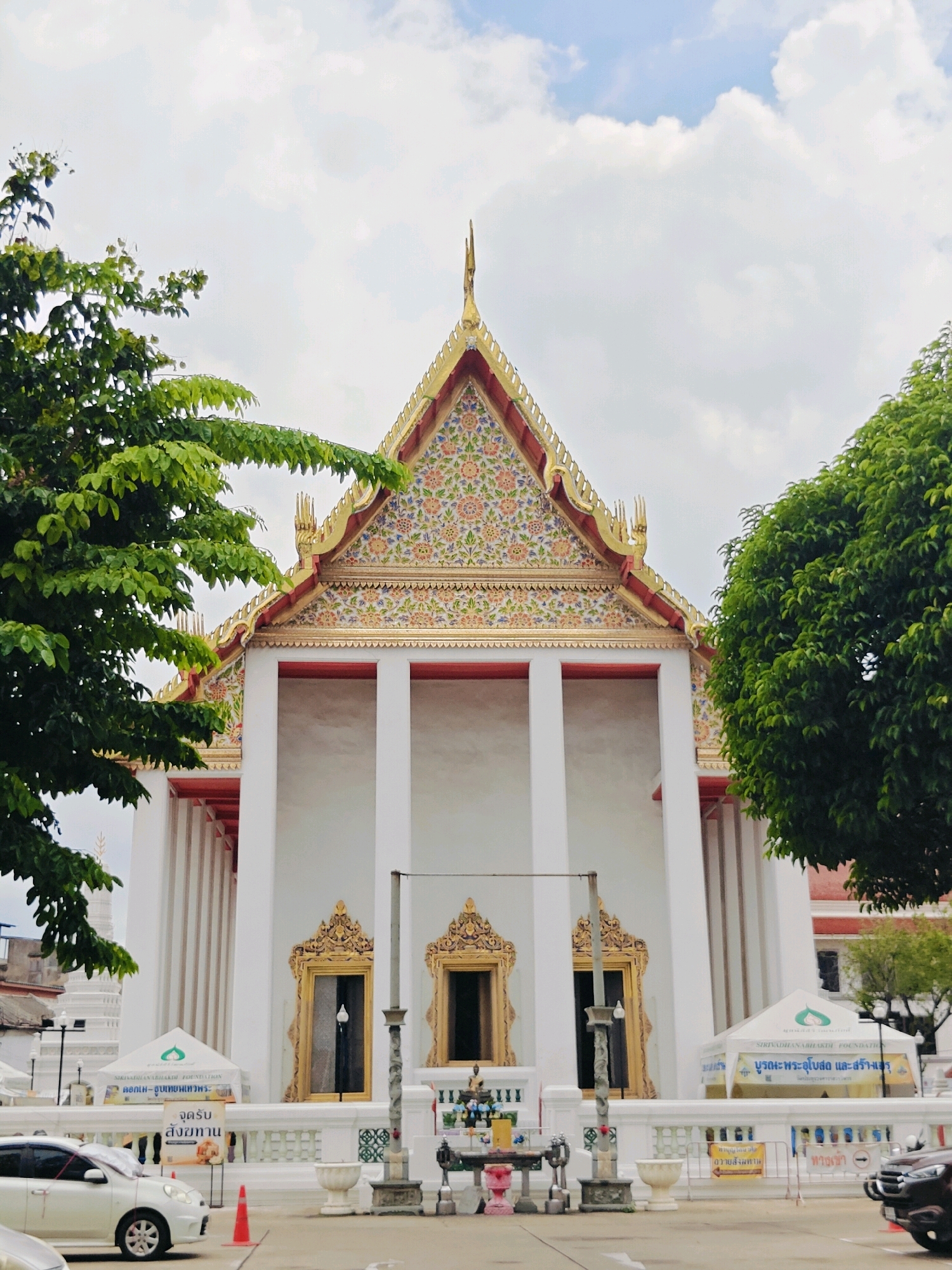 The image shows the exterior of a Thai temple, likely in Bangkok, Thailand, due to its architectural style. The temple has a prominent white facade with large columns and a decorative, gilded roof adorned with colorful floral patterns. In front of the temple, there are white tents with blue lettering, possibly for events or a foundation. Several cars are parked in the foreground, including a white sedan and a dark SUV. A small shrine with offerings is visible near the temple entrance. Green trees frame the sides of the temple, and a cloudy sky is visible above.