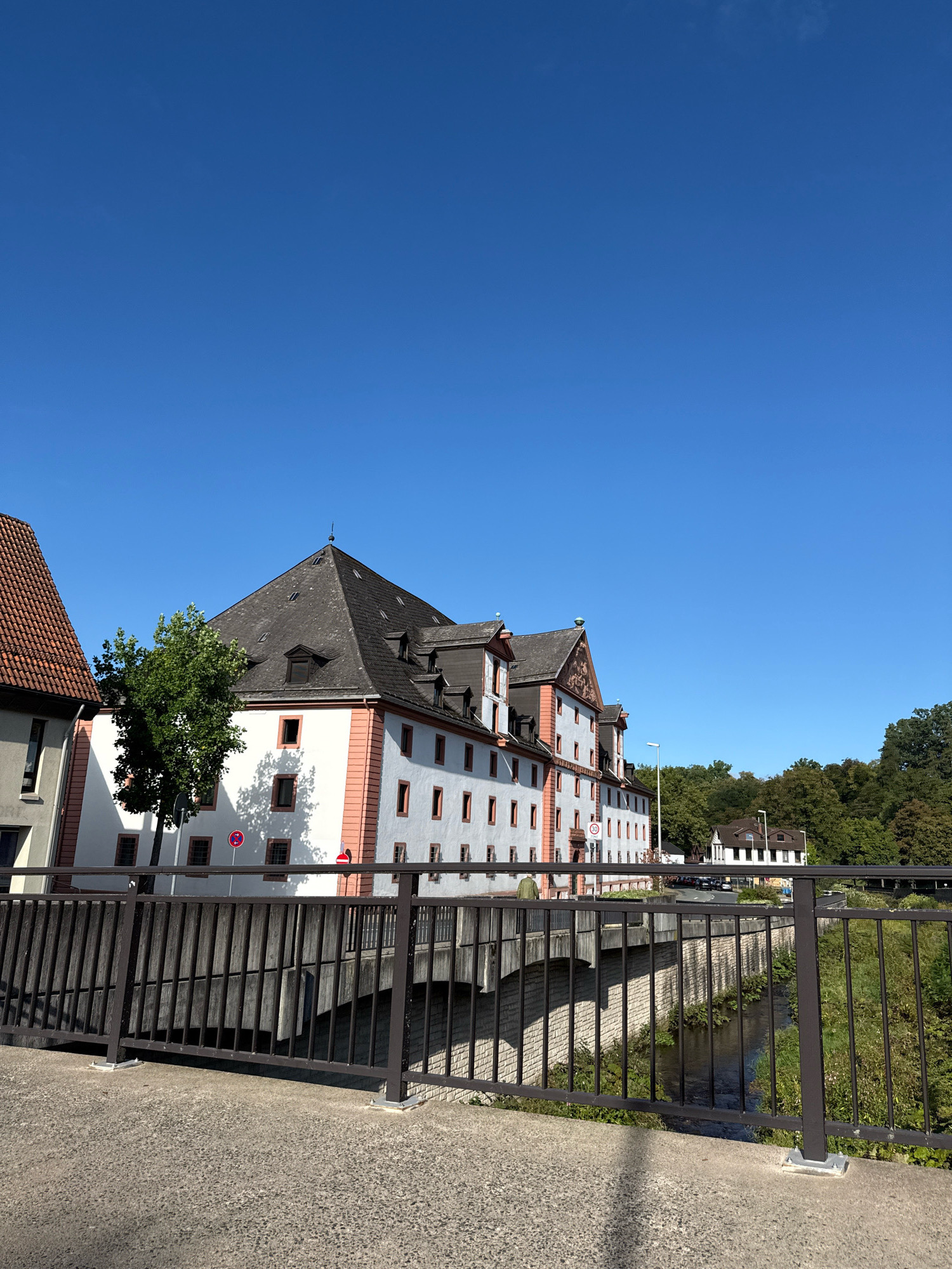 This image captures a sunny day scene in Osterode am Harz, Germany, featuring a historic building with a distinctive dark grey mansard roof and white walls accented with red trim. The building has numerous evenly spaced windows with dark frames. In the foreground, a metal railing with vertical bars borders a concrete bridge over a narrow, shallow river with overgrown banks. Adjacent to the main building, another building with a reddish-brown tiled roof is partially visible. A mature tree with green foliage stands to the left of the main building, casting a shadow on its facade. The sky is a clear, vibrant blue. In the distance, a smaller white building with a dark roof is visible, surrounded by dense green trees. The scene conveys a sense of a peaceful, well-preserved historical district.