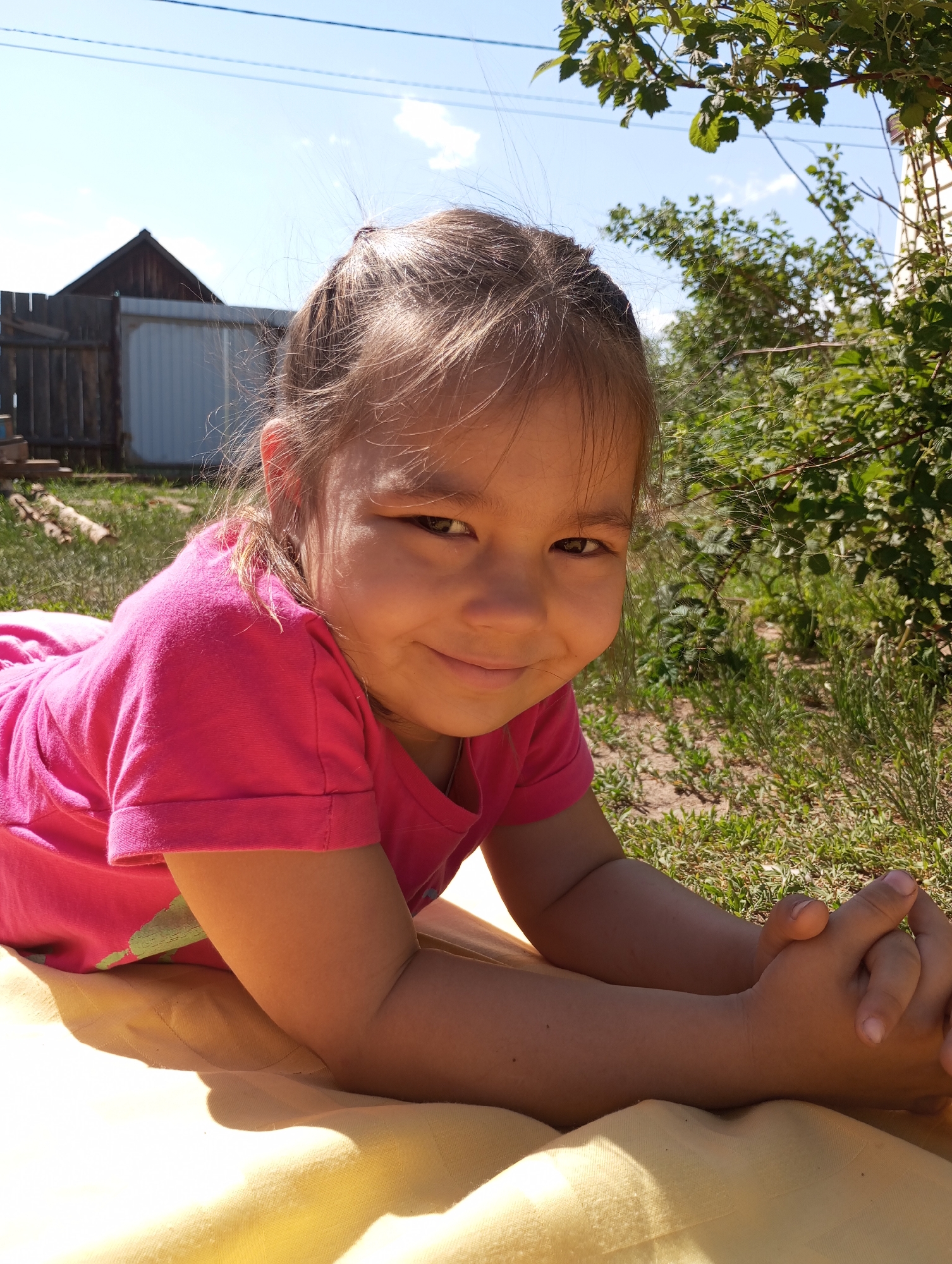 A young girl is lying on her stomach on a yellow blanket in a grassy area in the city of Буренка, Russia. She is wearing a bright pink t-shirt. Her hands are clasped together in front of her. Behind her is a wooden fence, a white garage, and some greenery. The sky is blue with a few clouds. The lighting suggests it is a sunny day.