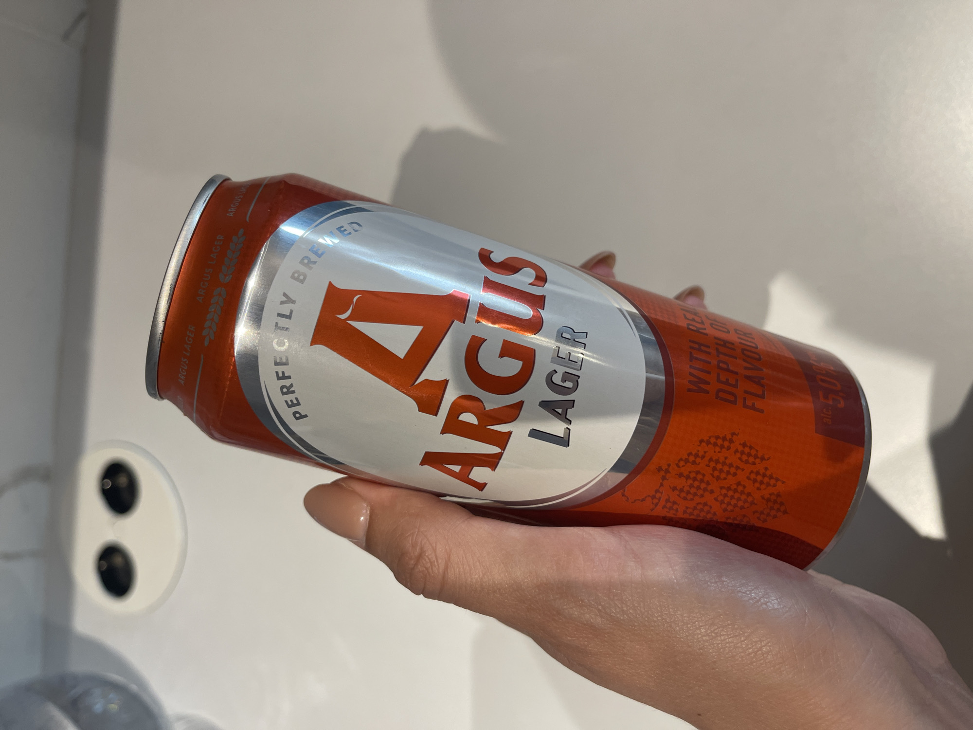 A hand holds a can of Argus Lager. The background is a plain white surface. The can is orange and silver with the "Argus" logo prominently displayed. The text "PERFECTLY BREWED" and "WITH REAL DEPTH OF FLAVOUR" are also visible. The can indicates it contains 500ml. The presence of "Argus Lager" suggests a European or possibly Bulgarian setting, but there are no definitive indicators of Sofia, Bulgaria.
