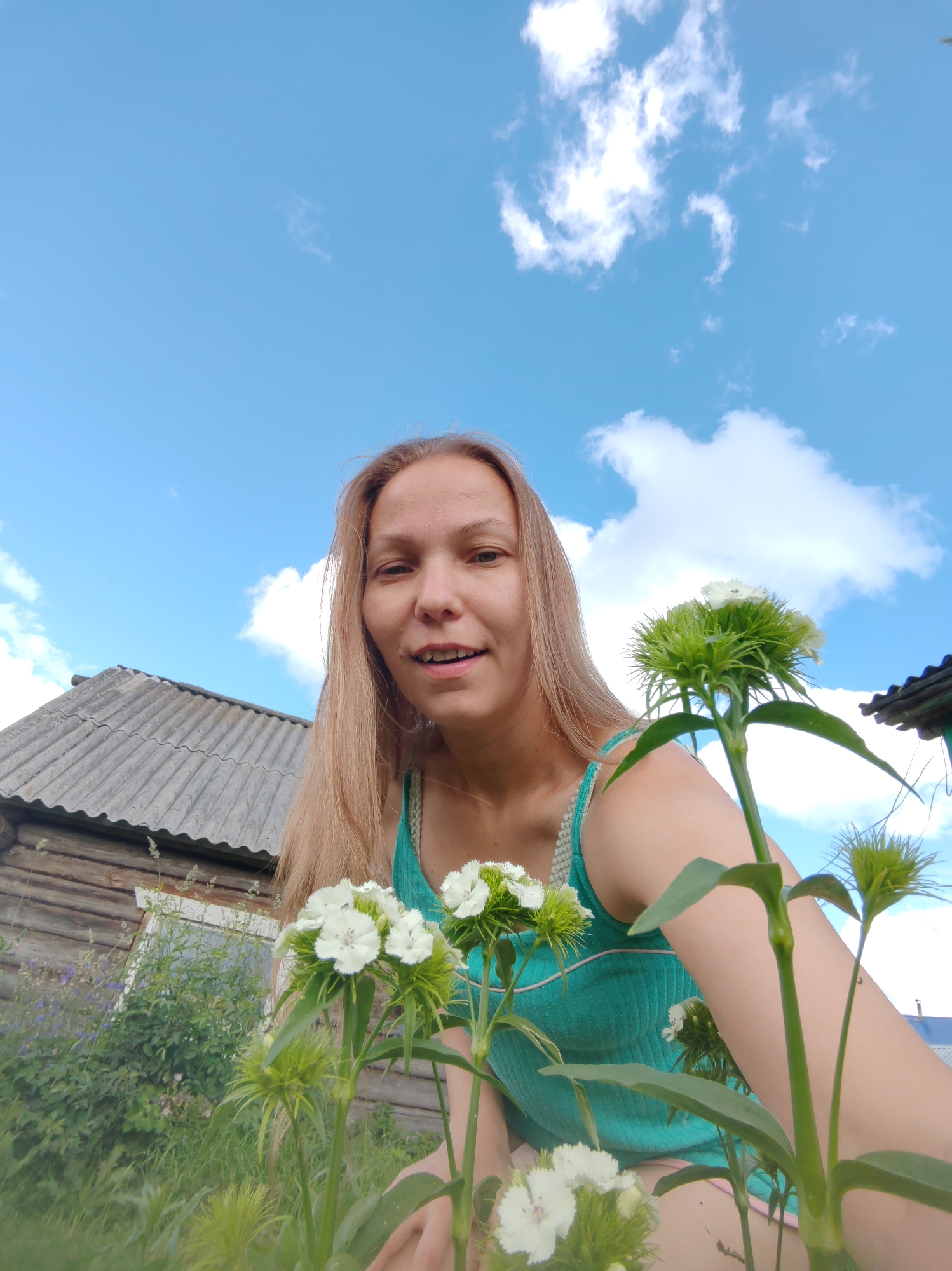 A young woman with long blonde hair is captured in a low-angle, outdoor, selfie-style shot. She is wearing a teal tank top and is positioned in the foreground, with a cluster of small white flowers in front of her. Behind her, a rustic wooden building with a corrugated roof is visible, suggesting a rural or semi-rural setting. The background consists of a bright blue sky with scattered white clouds. The overall atmosphere is casual and summery. The image appears to be taken in the village of Chukhlem, Russia.