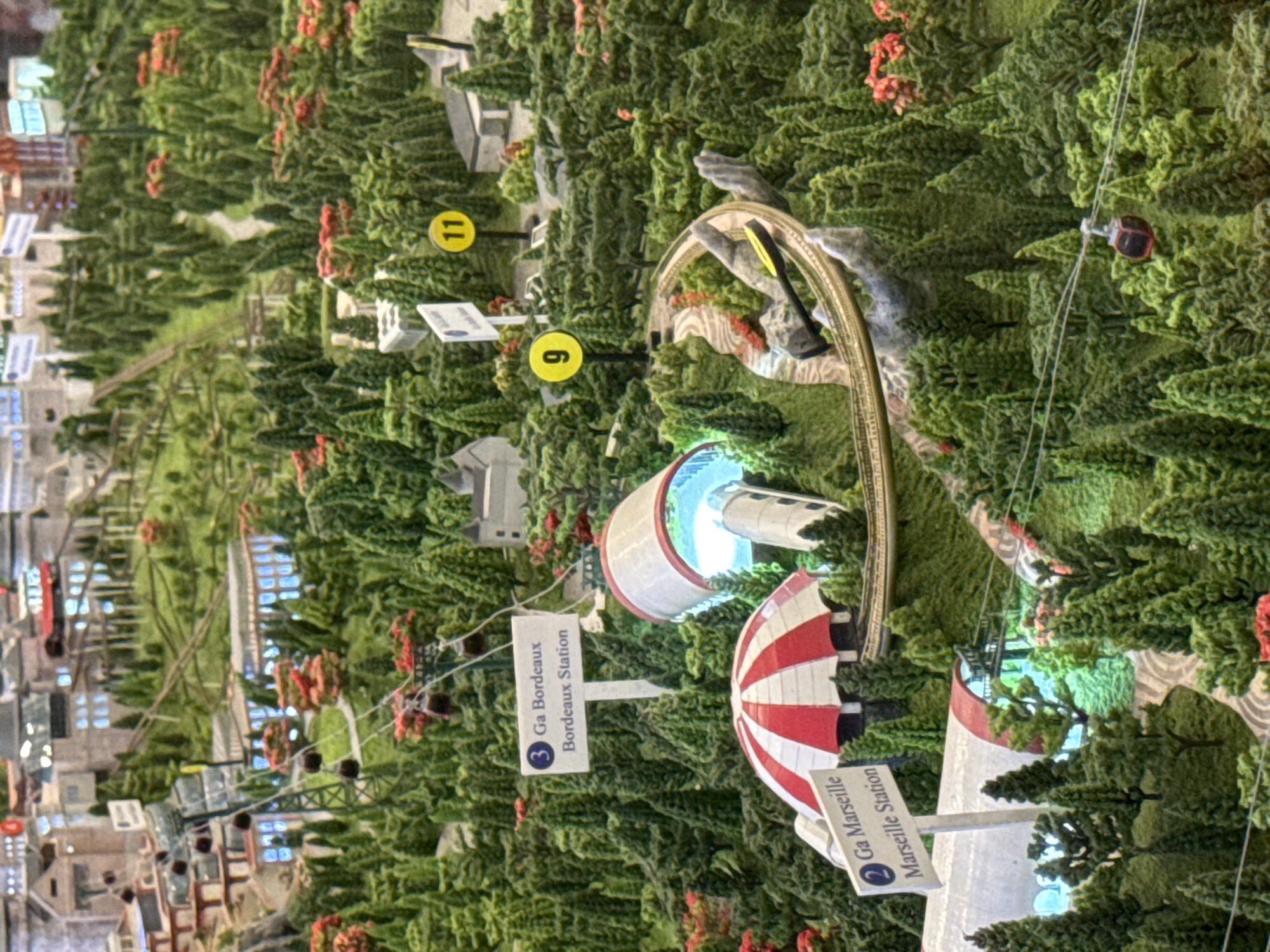 The image presents a miniature model representing a city, identifiable as Đà Nẵng, Vietnam, based on the included signage. The miniature features a detailed cityscape transitioning into a mountainous, forested region. Key identifiable structures and features include:
* **Built Environment:** A dense urban area with numerous buildings, varying in size and style, is depicted on the left side.
* **Transportation:** A cable car system runs from the city towards the mountainous region, with cars visible in the air. An elevated railway track snakes through the forested area.
* **Signage:** Signs with the labels "2 Ga Marseille Marseille Station" and "3 Ga Bordeaux Bordeaux Station" are visible, suggesting these are stations within the model. Numbered markers, "6" and "11", indicate additional points of interest.
* **Geographic Features:** A transition from a dense city landscape to a forested area with sculpted trees and a mountainous terrain. A sculpted hand holding a rail track is visible.
* **Lighting:** Internal lights illuminate specific structures and areas within the model, adding depth and contrast to the scene.
The scene is a diorama of a section of Đà Nẵng, exhibiting the city's juxtaposition of urban development and natural landscapes.