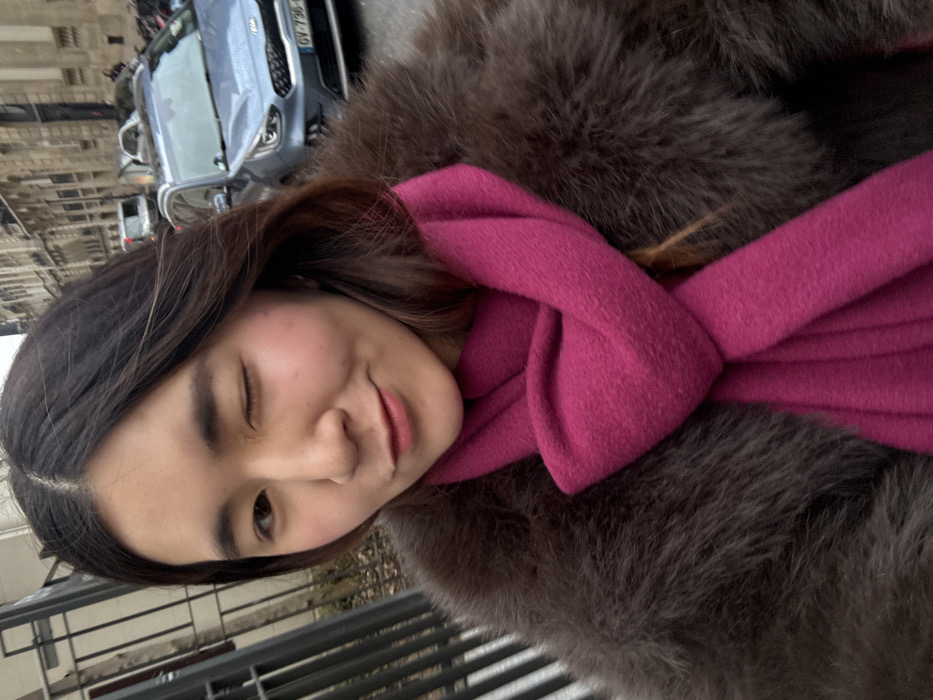 An East Asian female, estimated 20-30 years old, is depicted in a close-up selfie taken outdoors on a street in Paris, France. She has long, dark brown hair with a center part and is wearing a dark brown, fur-textured coat with a bright magenta scarf wrapped around her neck. Her left eye is closed in a wink, while her right eye is open, and her lips form a subtle smile. To her immediate left, a dark metal fence is partially visible. The background to her right and behind her features an urban street scene with a light blue-grey Kia Niro SUV prominently displaying a French license plate, `GV-736-DR`. Multiple other vehicles are visible further down the street. The street is lined with multi-story, light-colored stone buildings exhibiting classic Parisian architectural styles, all under an overcast sky.